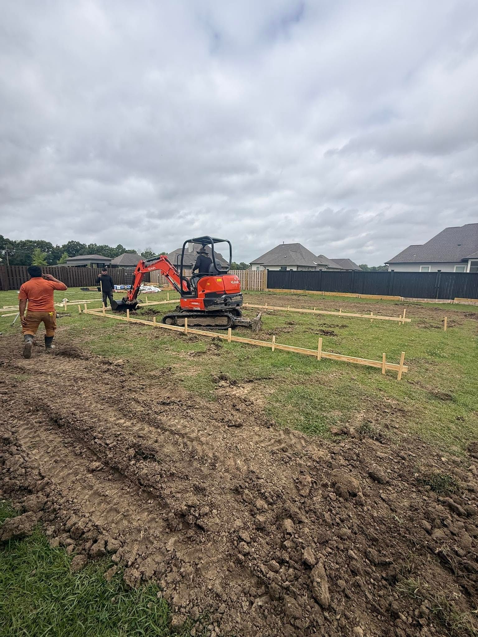 Construction workers operating an orange excavator in a muddy field near houses under cloudy skies