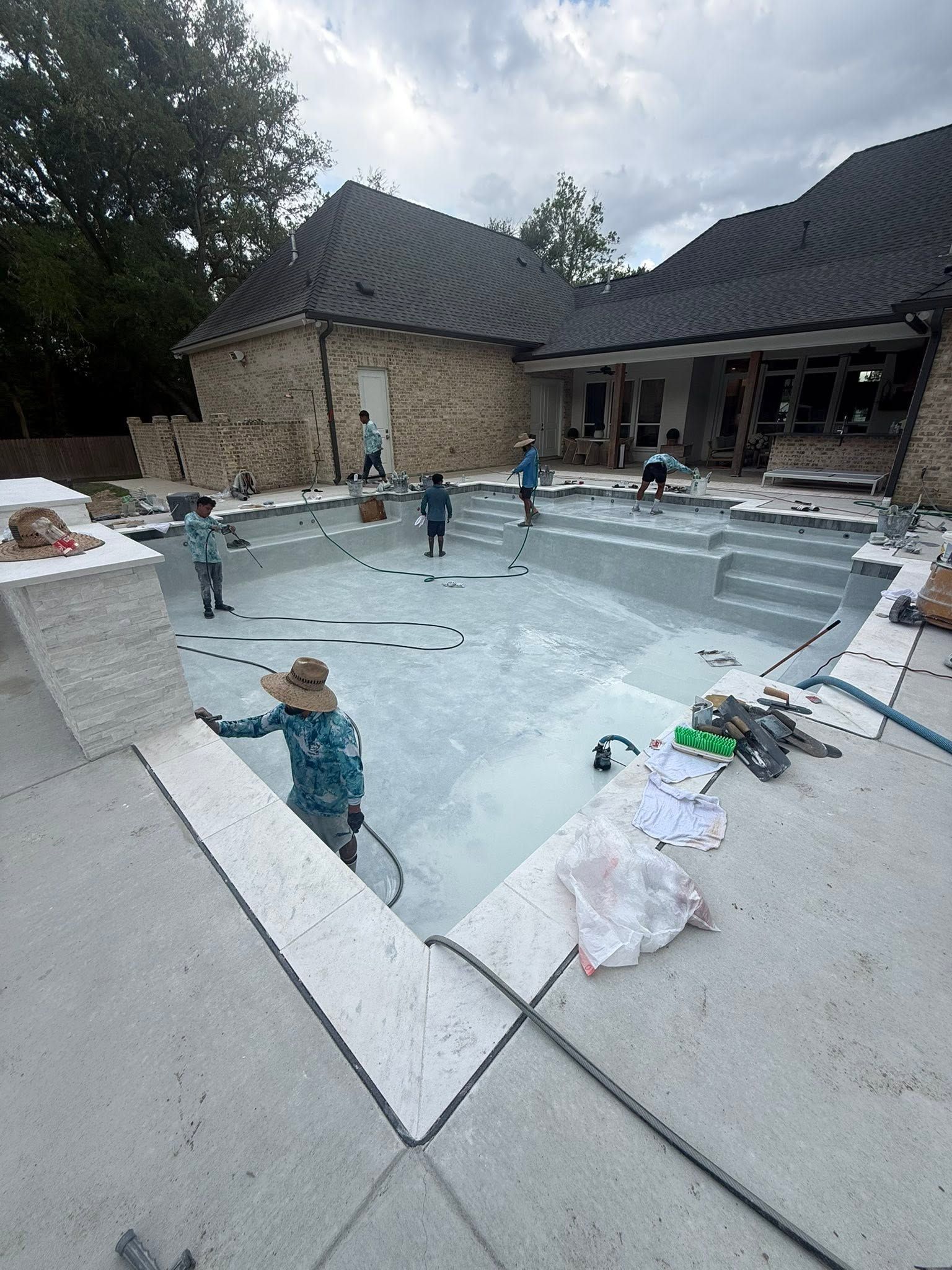 Partially built backyard pool with workers, blue water, and a stone house in the background