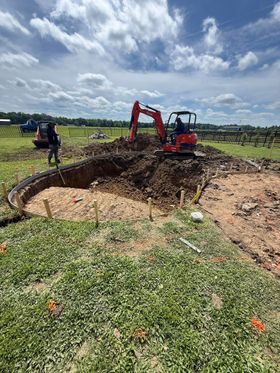 Excavator digging a large circular muddy pit in a grassy field under a cloudy sky