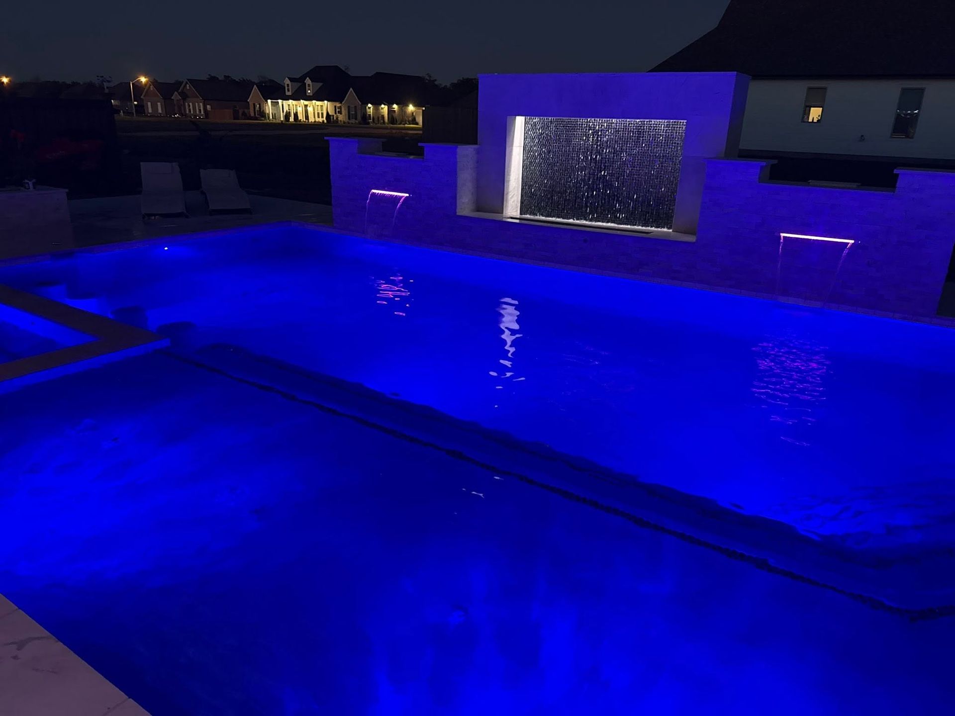 Blue-lit swimming pool at night with a lit waterfall feature and deck seating area