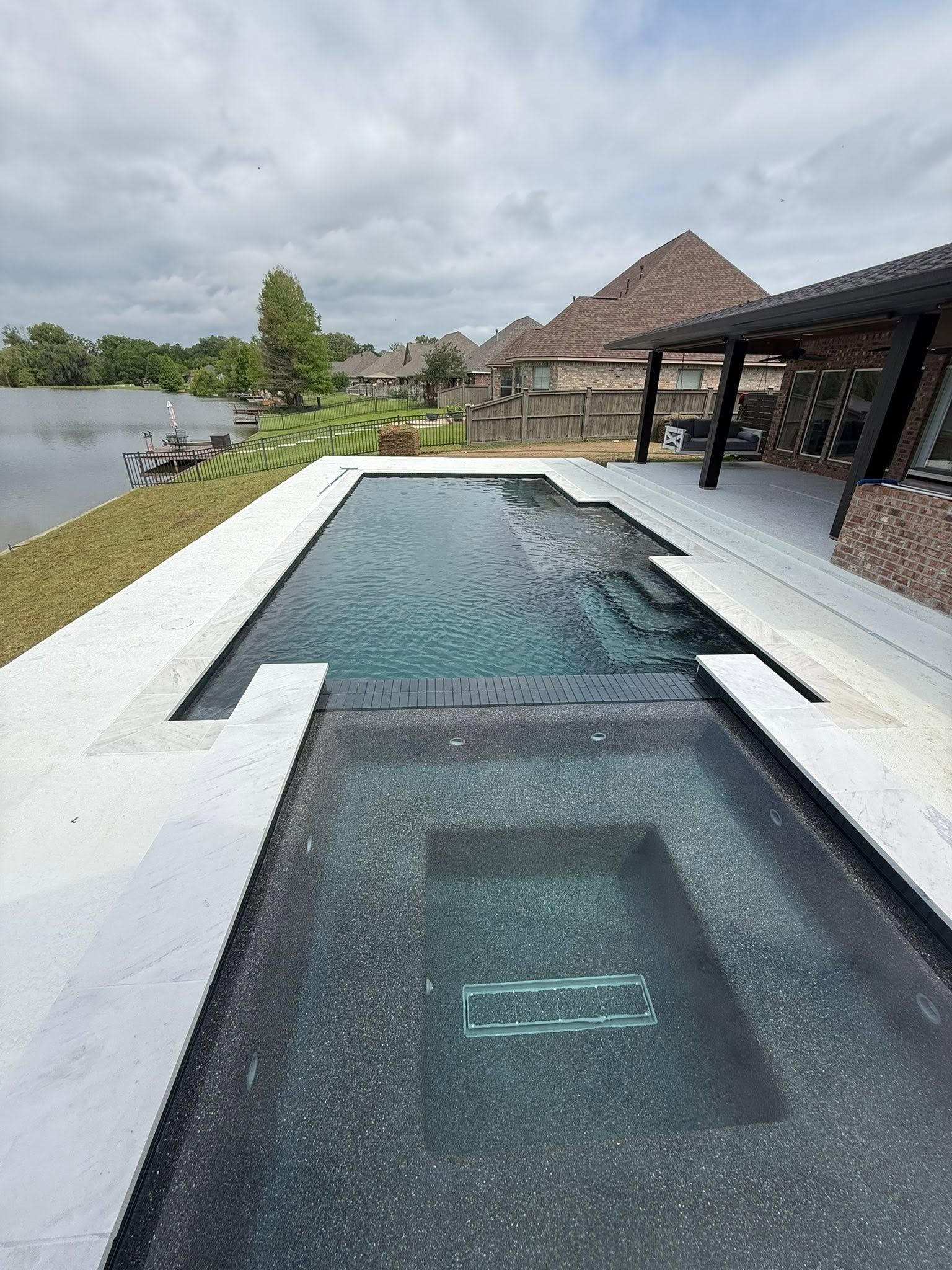Narrow outdoor swimming pool beside a lakeside patio with a thatched-roof pavilion and cloudy sky