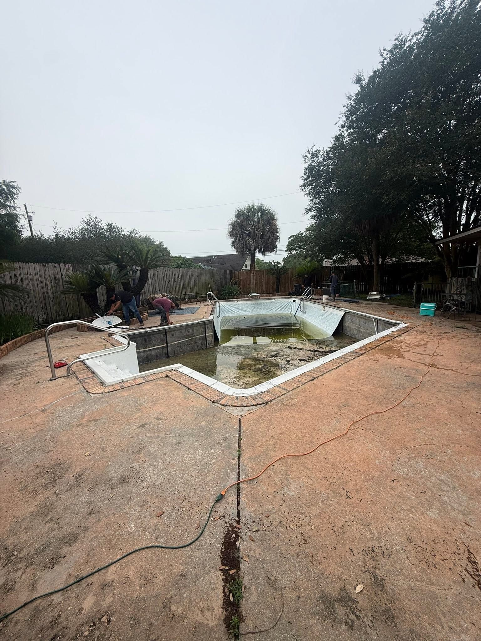 Broken concrete walkway around an empty, rectangular pool in a tree-lined outdoor area