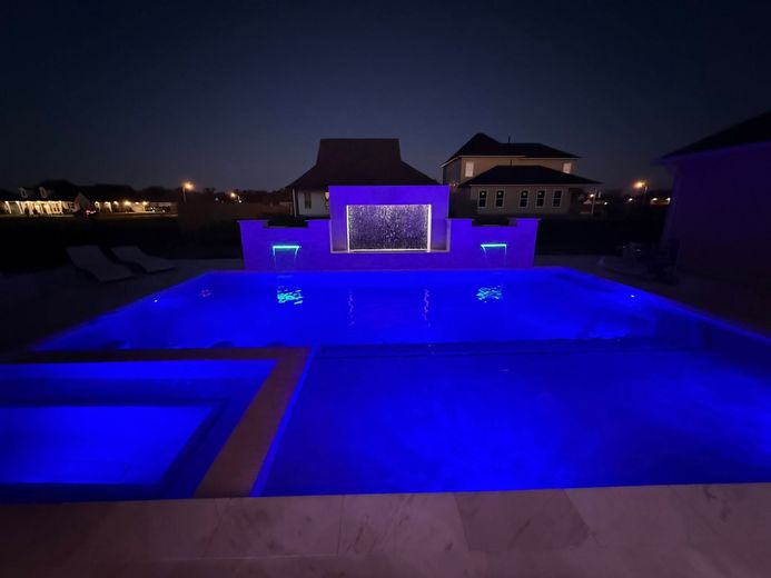 Blue-lit swimming pool at night with a lit waterfall feature and houses in the background