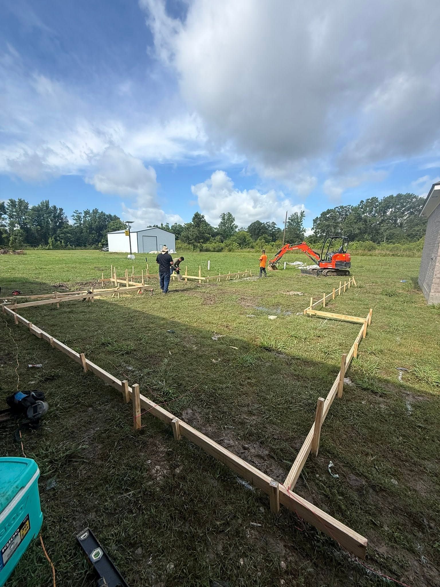 Workers marking out a foundation on a grassy lot, with an excavator and white building in the background