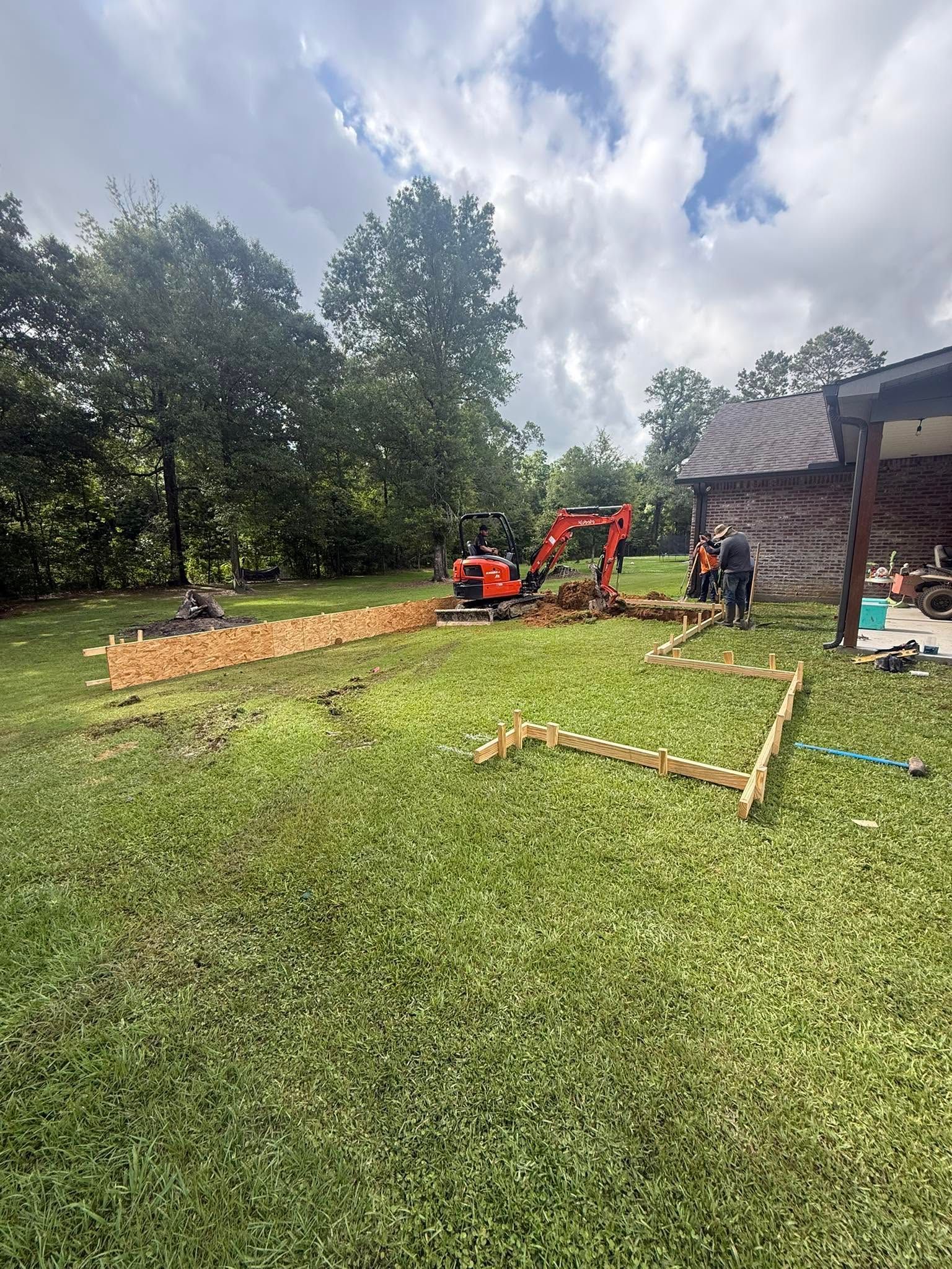 Construction workers framing a foundation on a grassy lot beside a brick house, with an orange mini excavator.