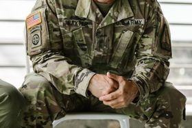 Soldier in camouflage uniform sitting with hands clasped, American flag patch visible on sleeve.