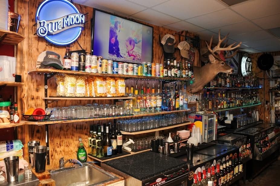 Cozy bar interior with a lit Blue Moon sign, shelves of bottles, and deer antlers mounted above the counter