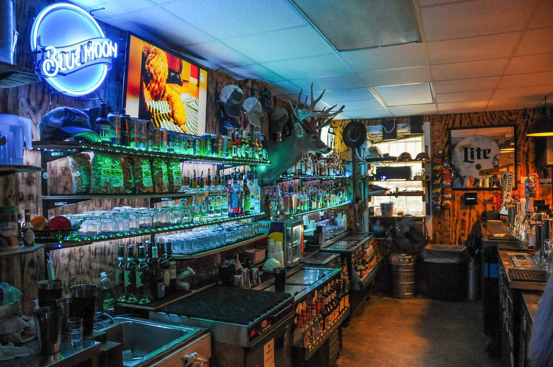 Neon-lit bar interior with shelves of bottles, posters, and a long counter under blue and orange lighting