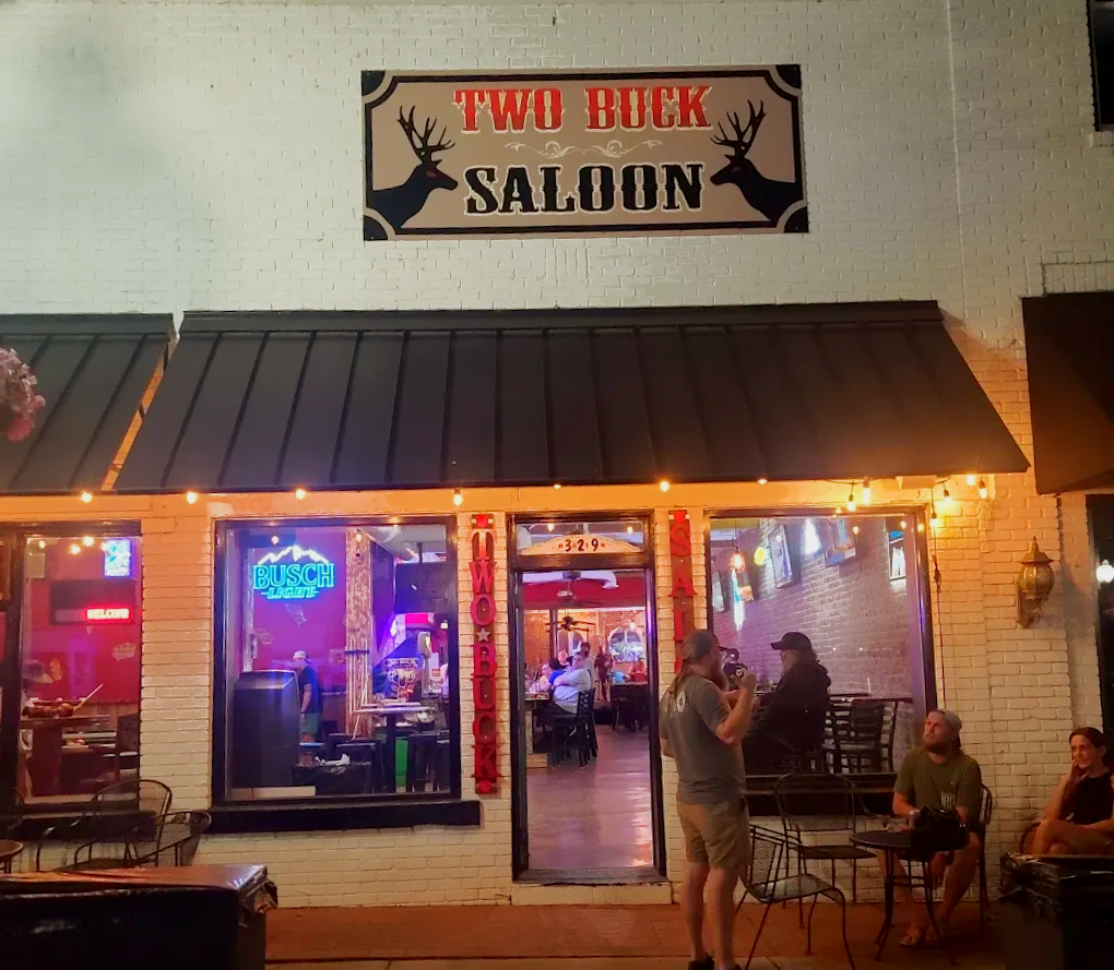 Two Buck Saloon storefront at night, lit by neon and string lights, with patrons outside and inside.
