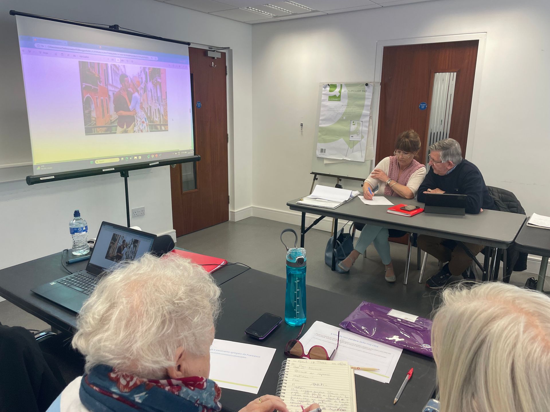 People in a meeting room, two at a table with papers, watching a presentation on a screen.