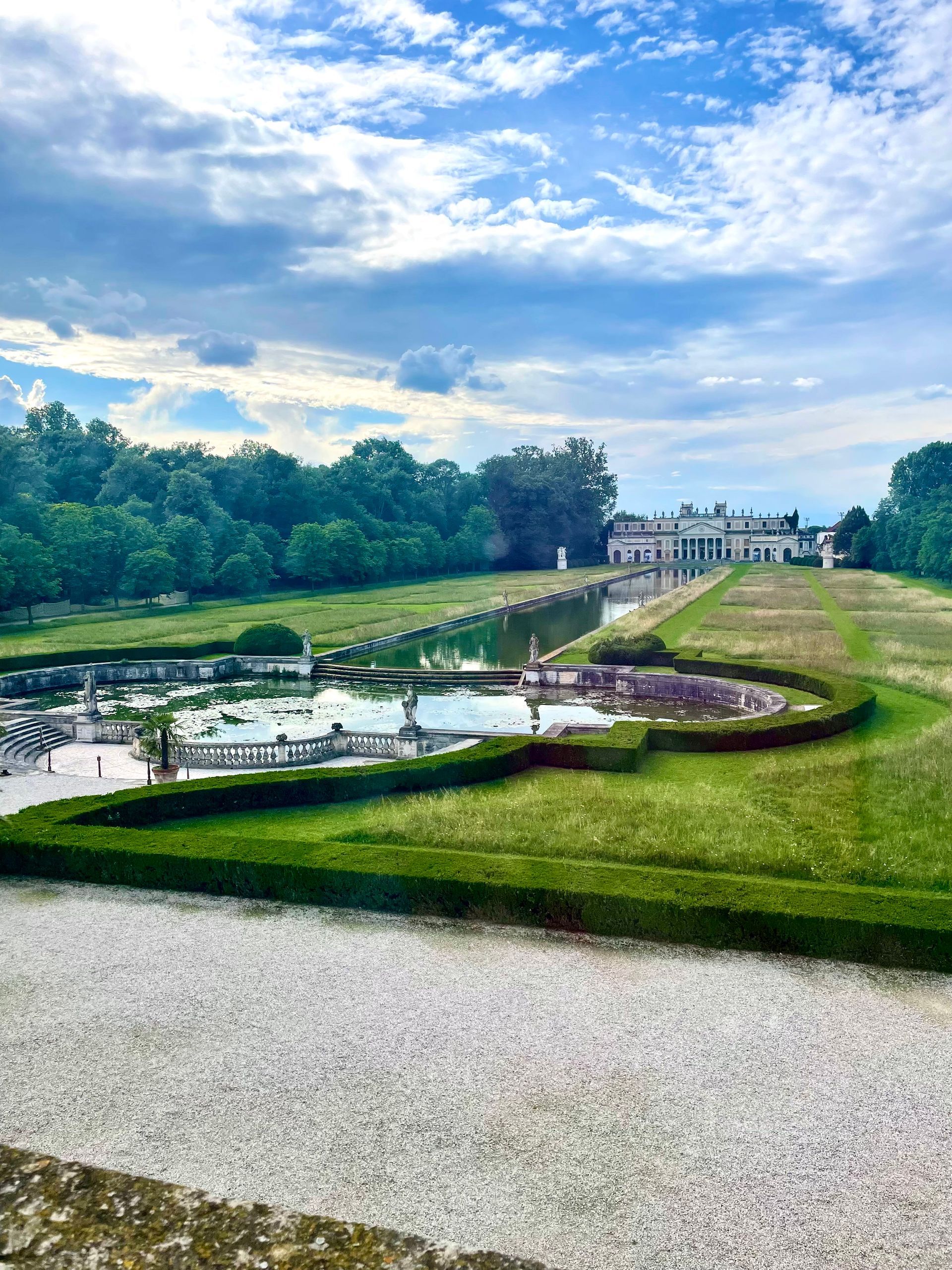 A mansion overlooks a formal garden with reflecting pools under a cloudy sky. Green lawn, trees.