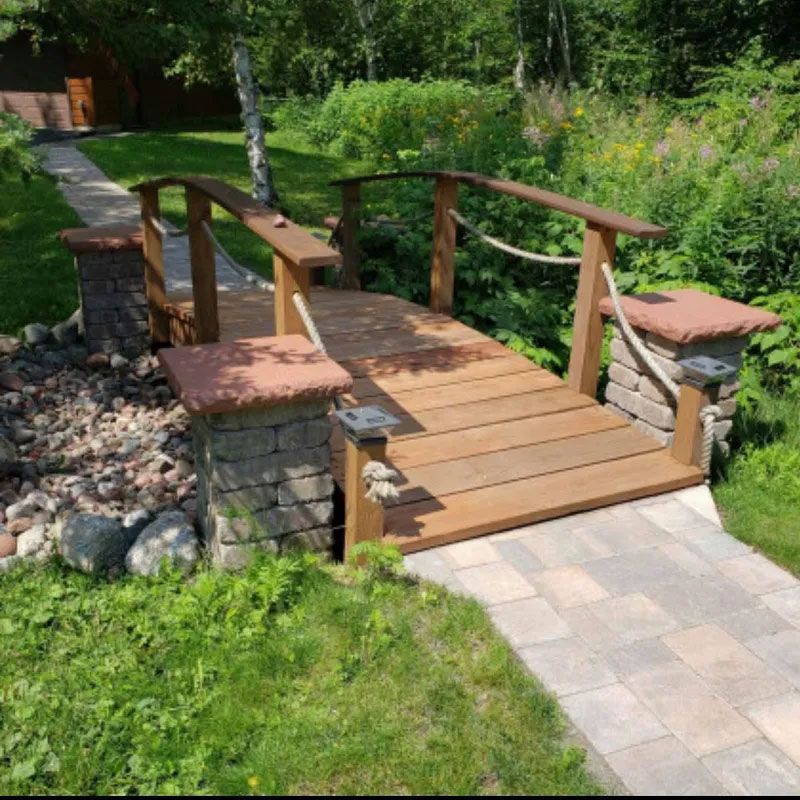 Small wooden bridge with stone supports over a garden path, surrounded by greenery and flowers
