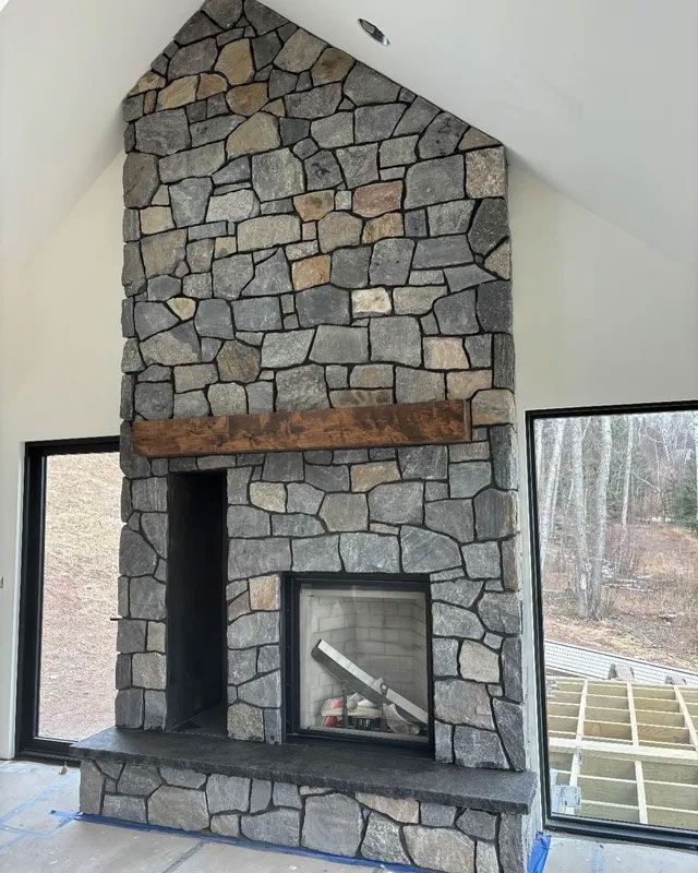 Stone fireplace with black firebox in a bright room with tall windows and a wooden mantel