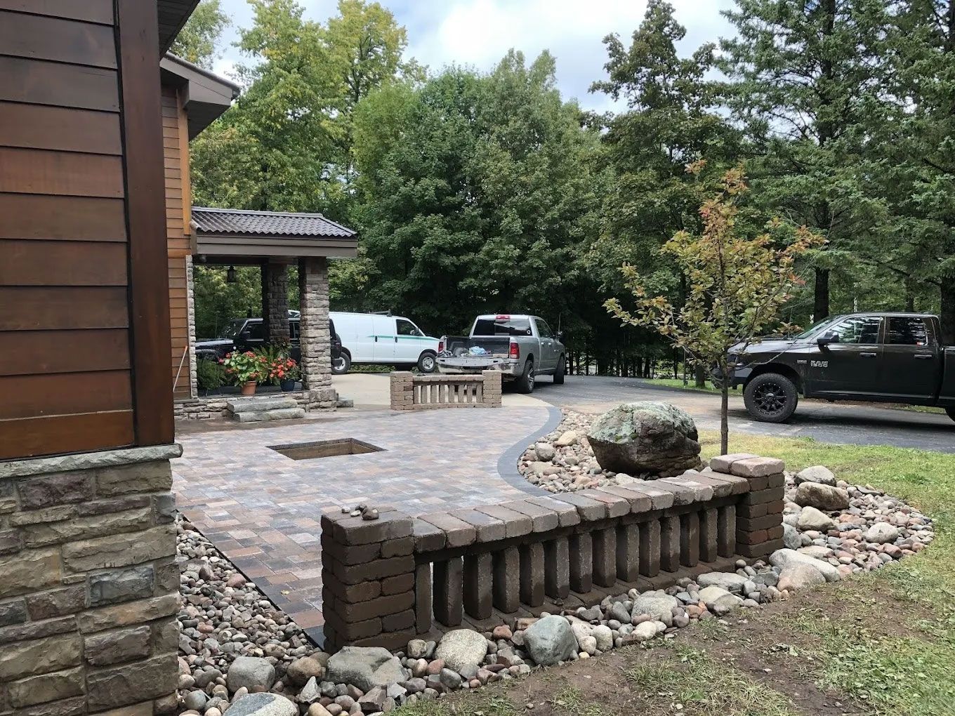 Stone patio with low brick walls, large rock, and parked vehicles beside a house under trees