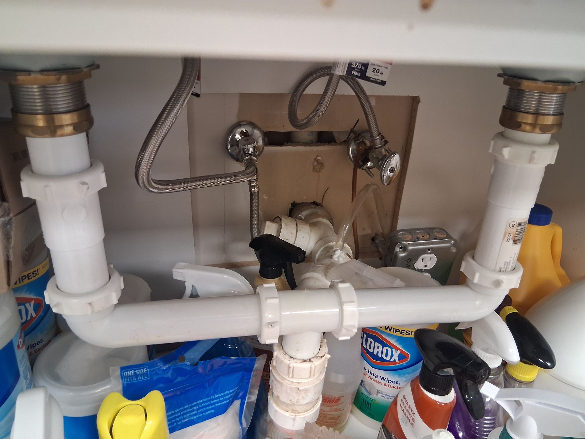Under-sink plumbing with white PVC pipes, valves, and cleaning supplies in a cabinet.