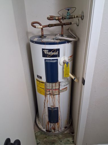 Old rusted water heater in a narrow utility closet with copper pipes and a warning label