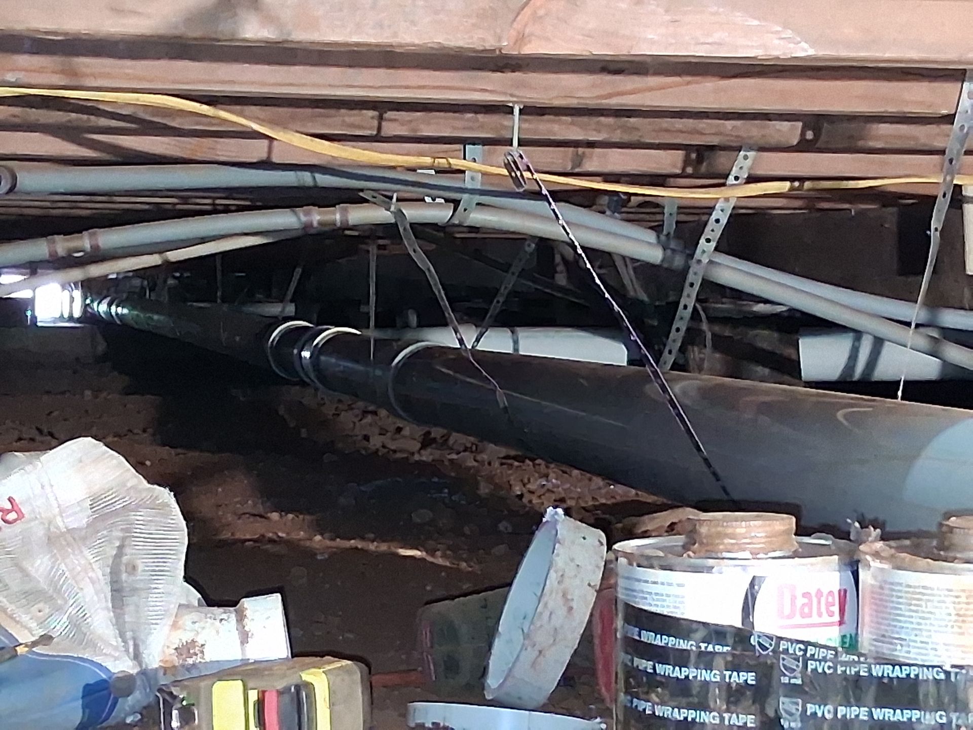 Cluttered crawlspace with exposed pipes, cables, and stacked cans under a wooden floor
