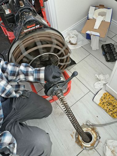 Person in plaid shirt and gloves working on a drain with a long snake in a tiled room.