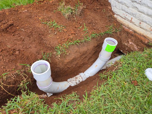PVC drain pipes installed in a dirt trench beside a brick wall and grass