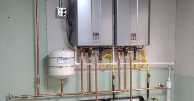 Utility room with wall-mounted boilers, copper pipes, and a white expansion tank on a pale green wall
