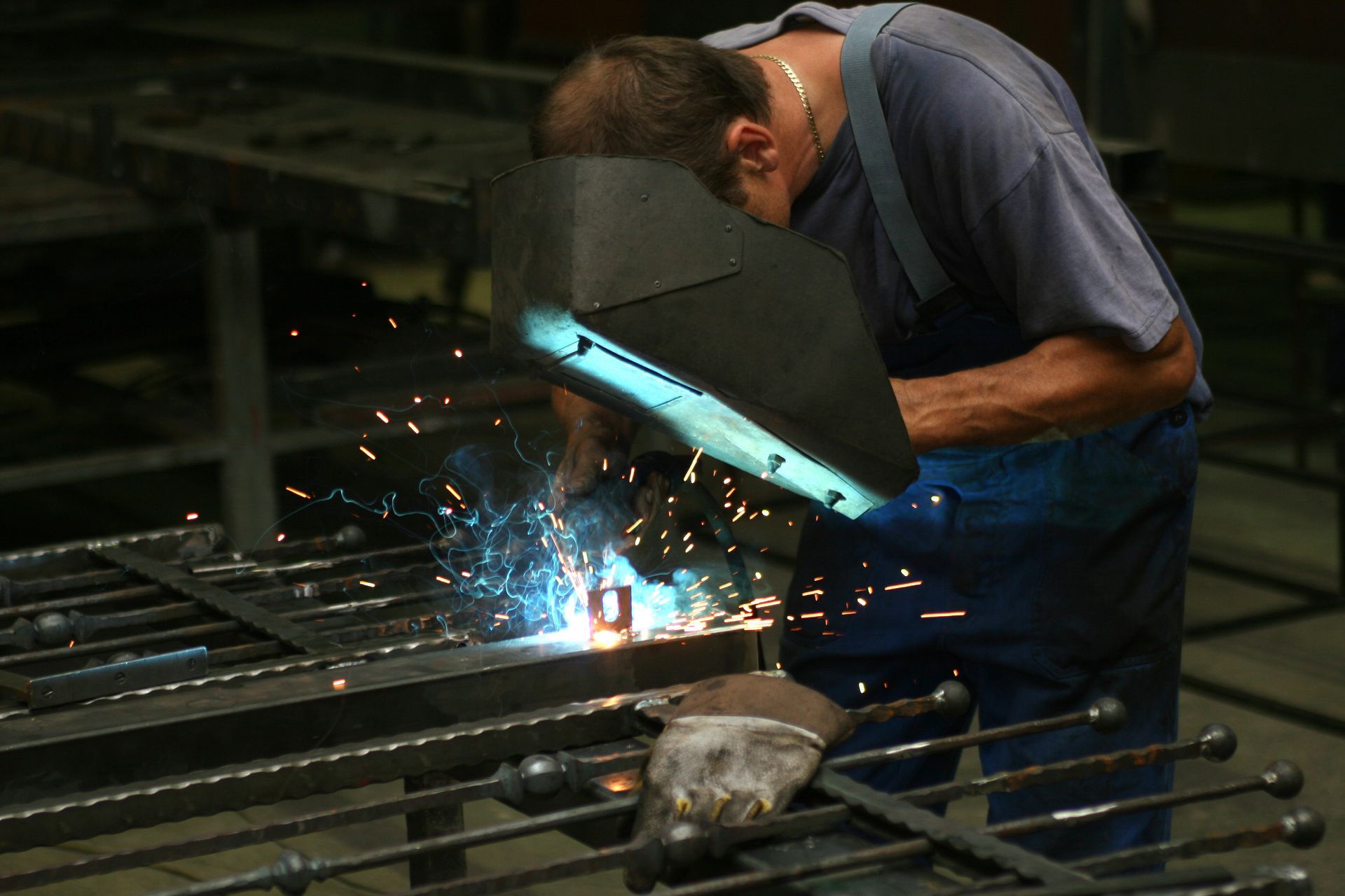 A person wearing a welding helmet performs arc welding on a metal gate in a workshop, creating bright sparks.