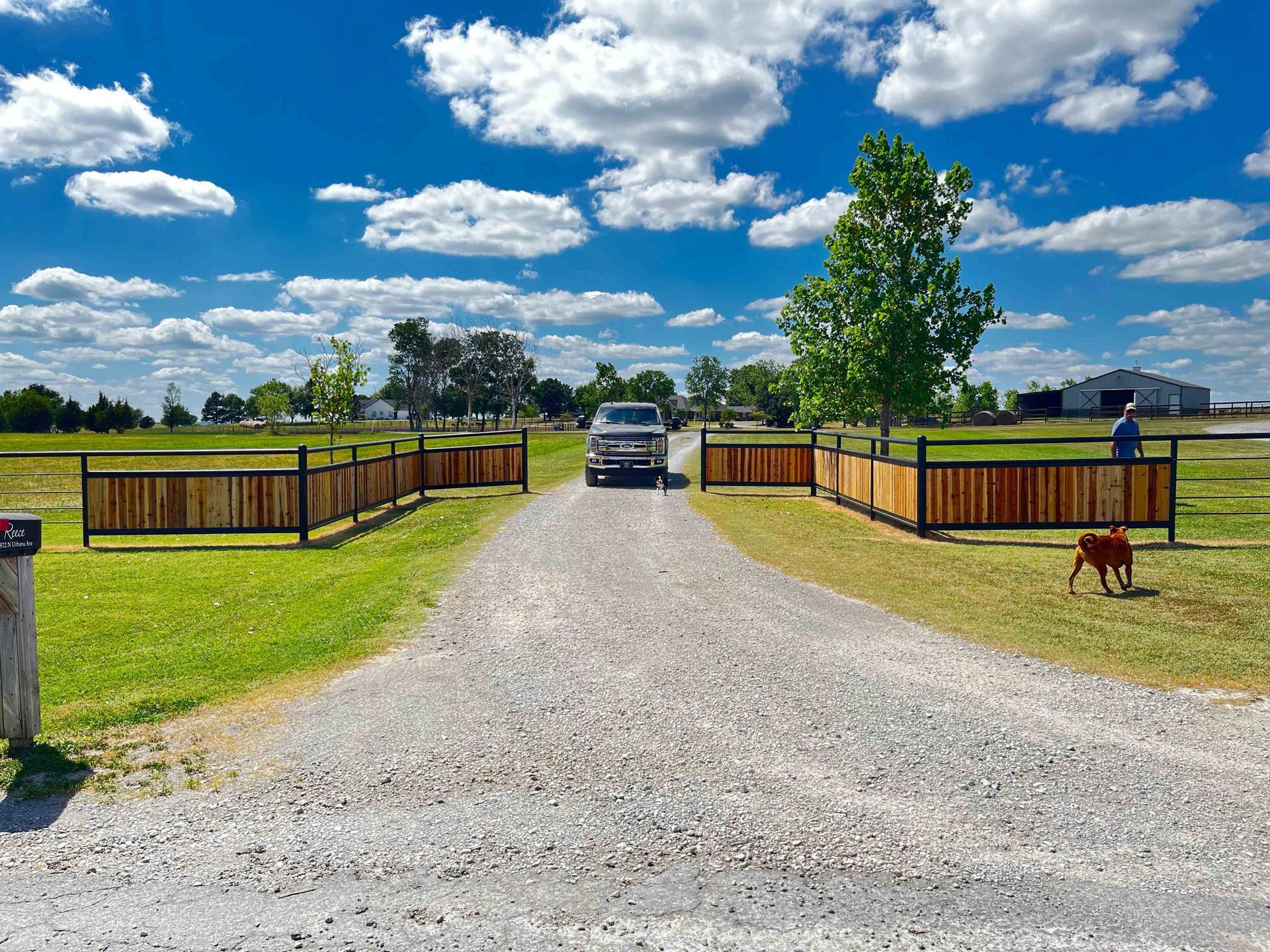 Gravel driveway to a farmhouse with fenced yards under a bright blue sky, car parked ahead.