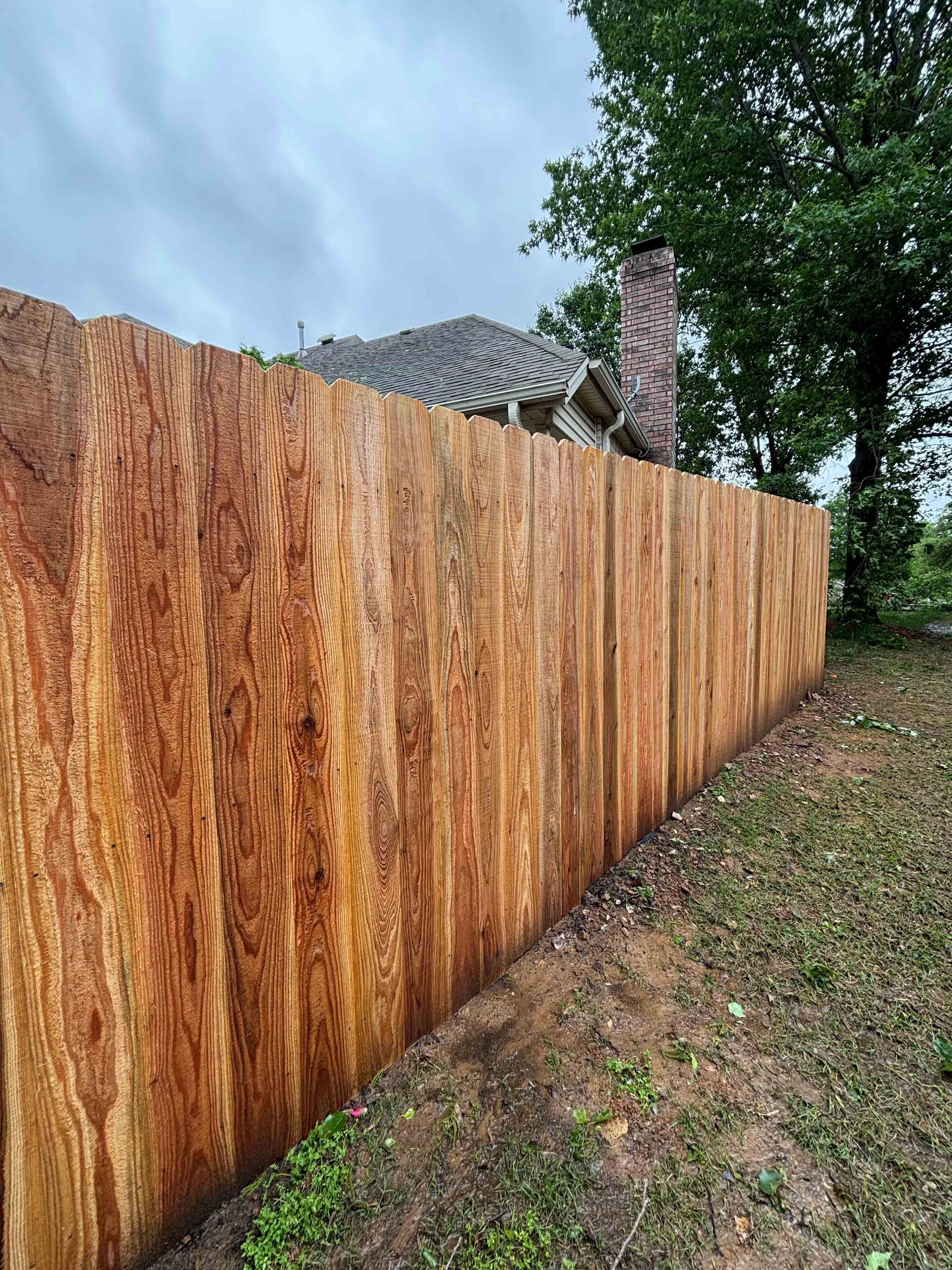 Tall rust-colored wooden fence beside a grassy yard under a cloudy sky, with a house behind it.