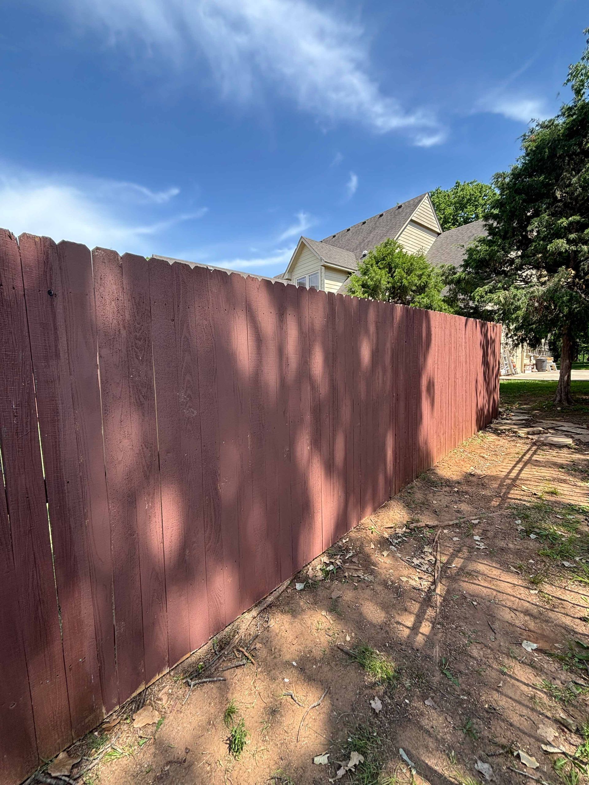 Weathered red fence beside a dirt yard, with a house and tree under a blue sky