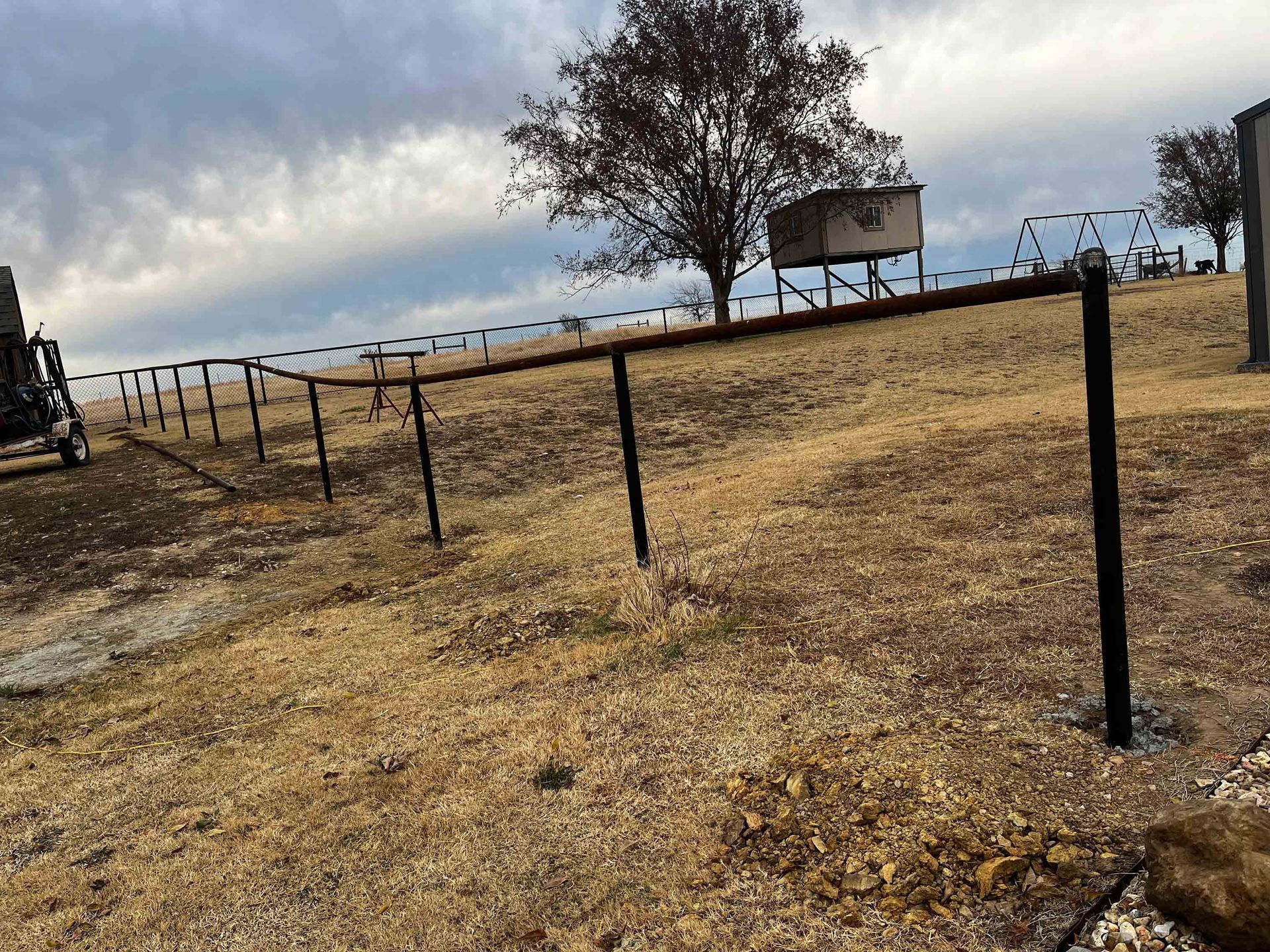 Dry grassy field with black fence posts leading to a small elevated lookout under a cloudy sky.