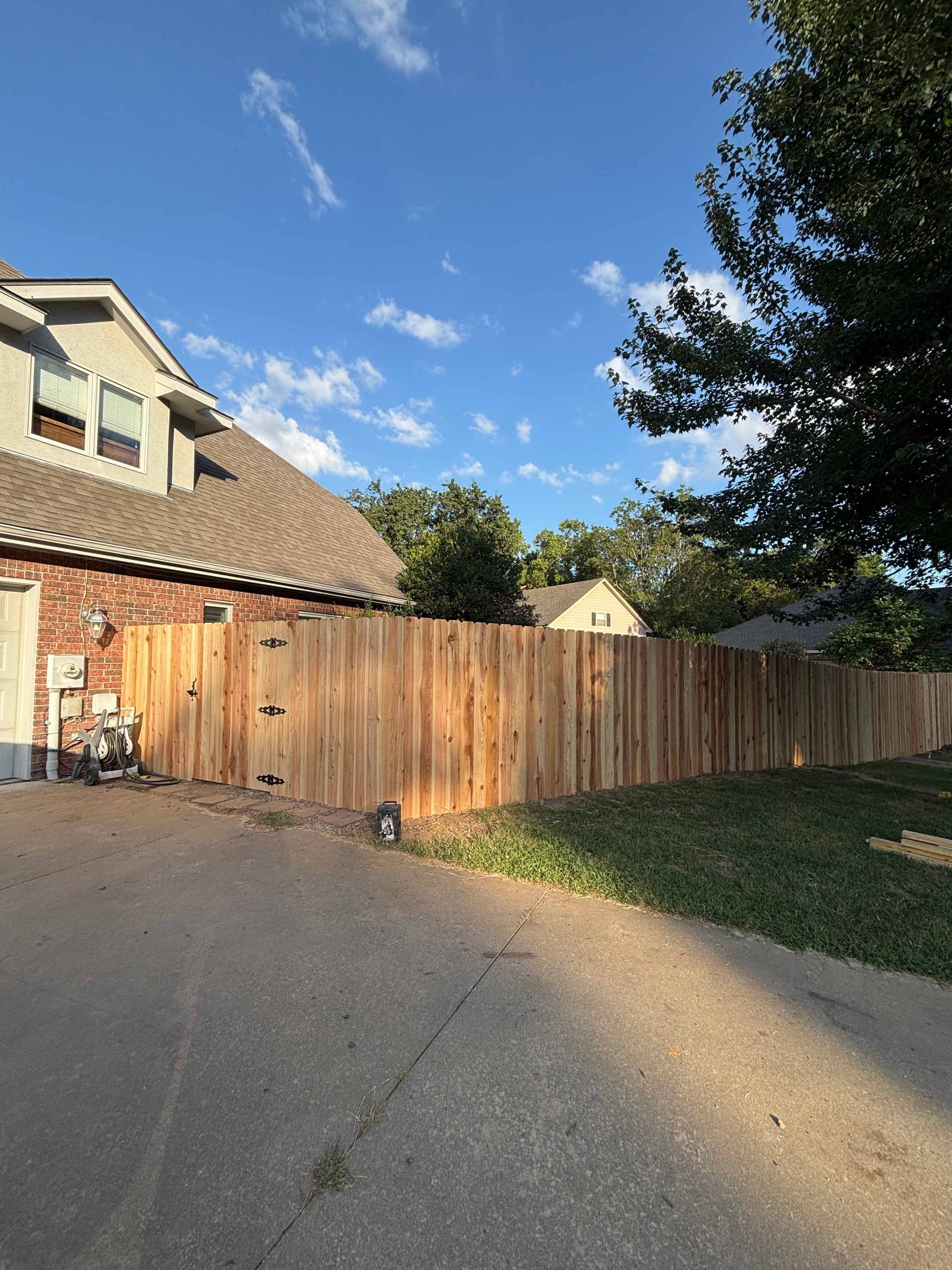 Wooden privacy fence beside a driveway and brick house under a blue sky with clouds.