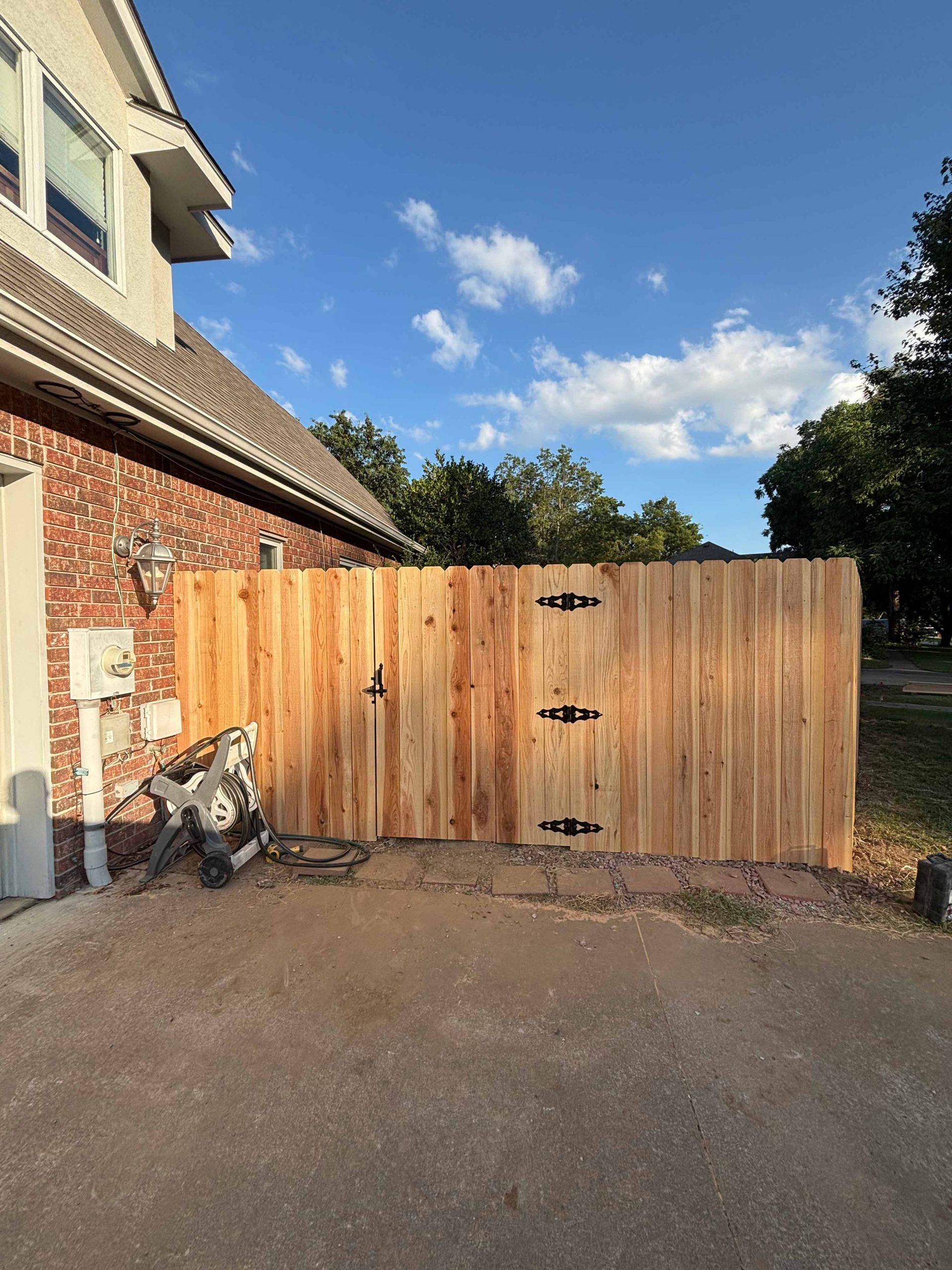 Wooden driveway gate beside a brick house under a blue sky with clouds