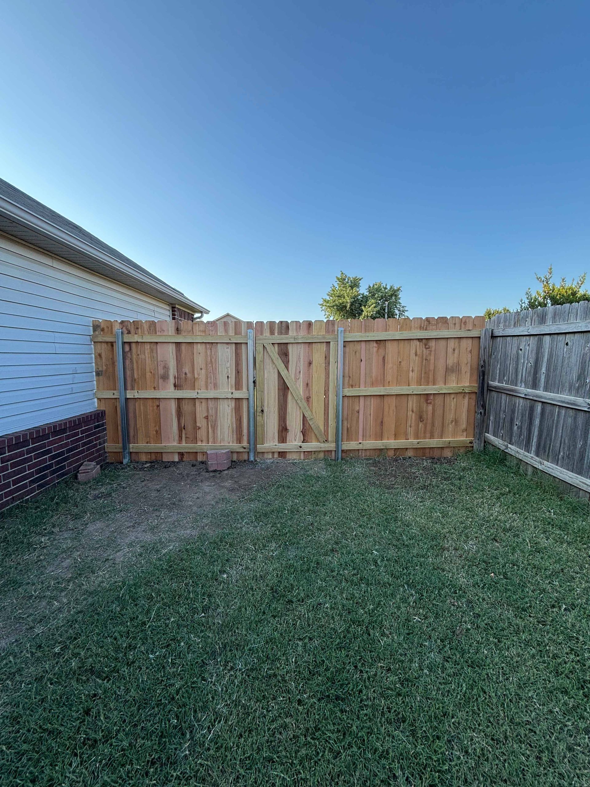 Backyard with a wooden gate fence between a white house and a weathered privacy fence under a clear blue sky