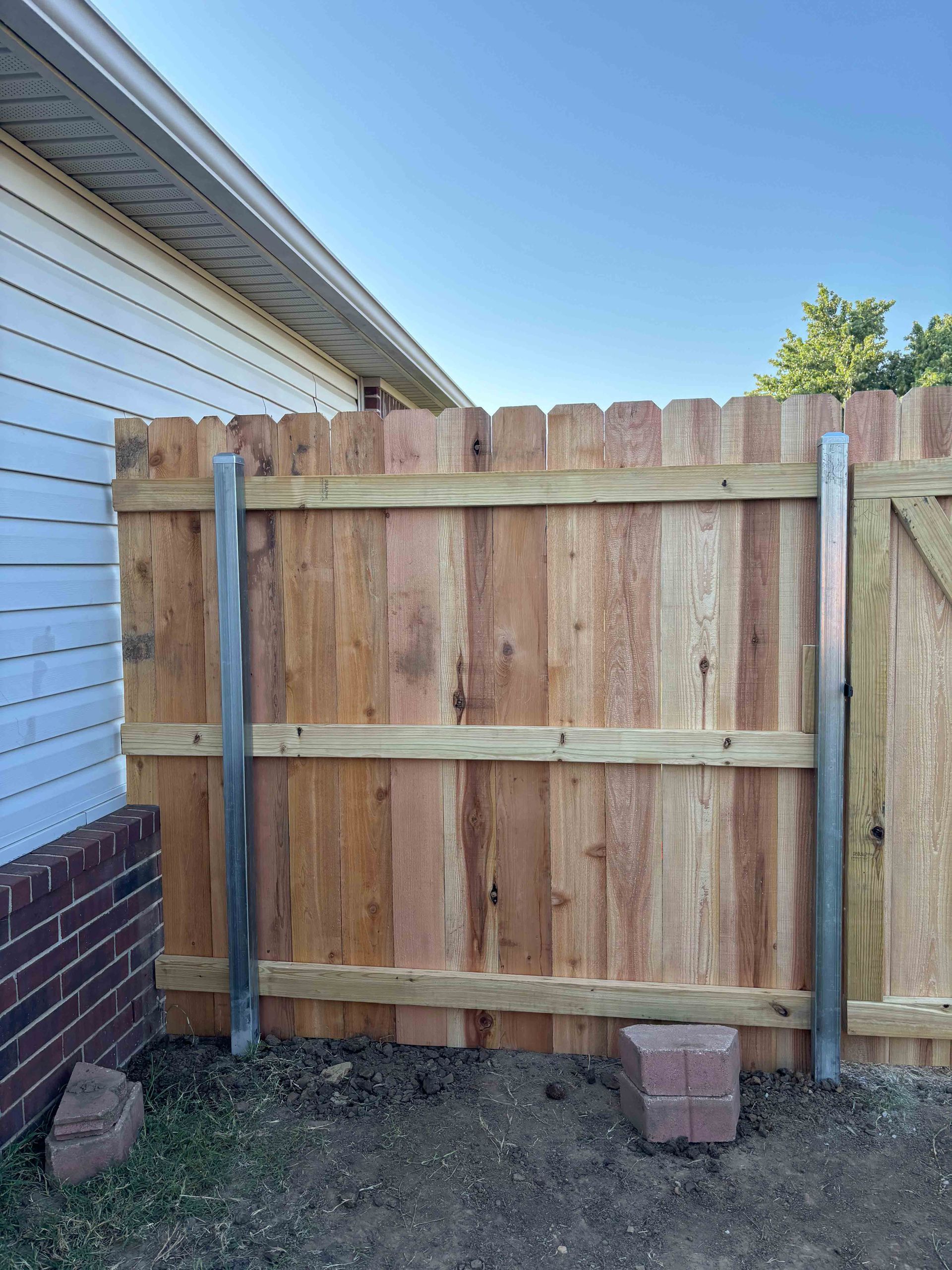 Wooden backyard fence gate between metal posts beside a house, with bricks at the base