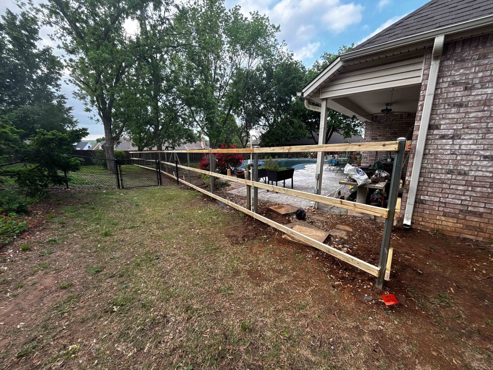 Wooden railing under construction beside a brick house, with tools and materials on a yard slope.