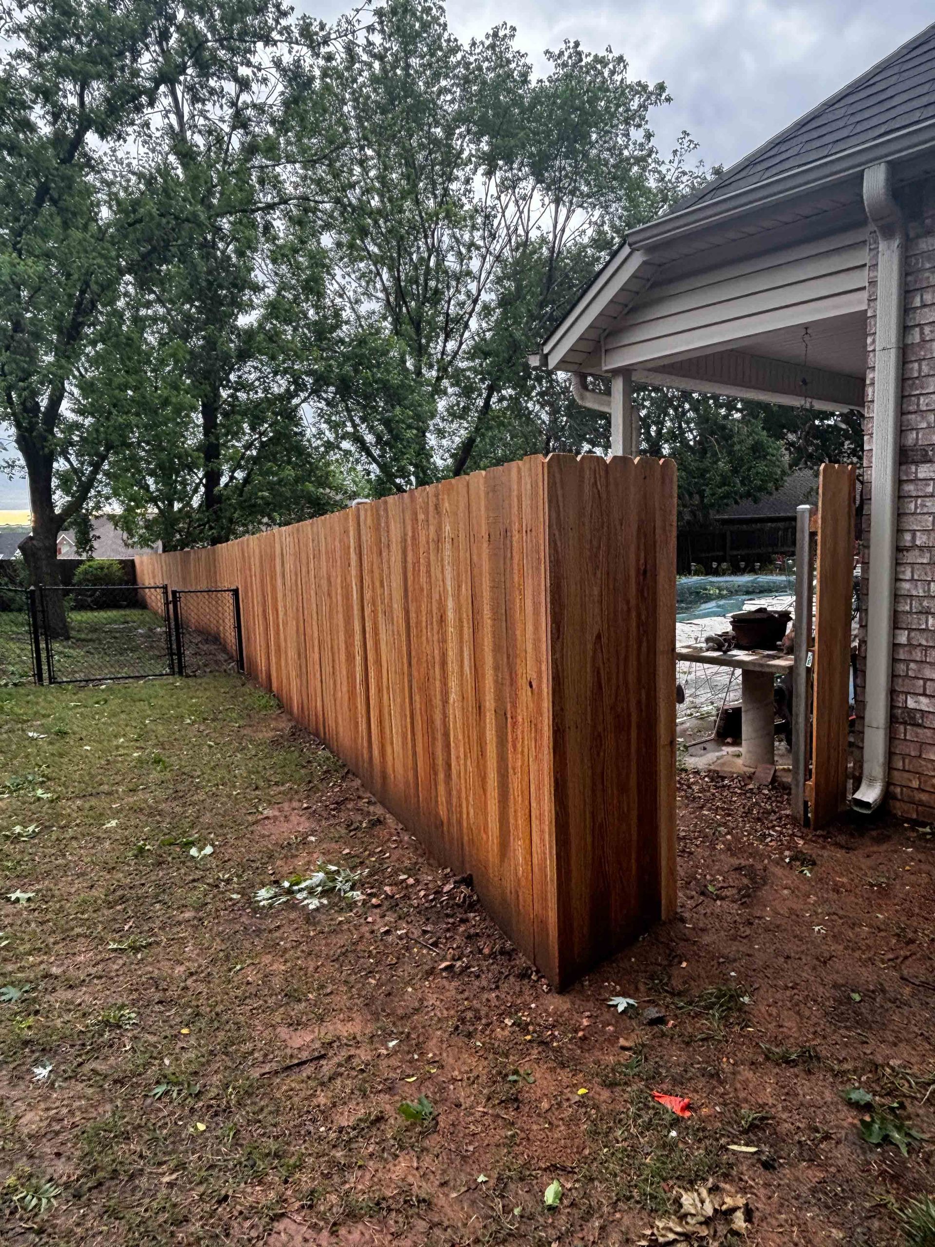 Wooden privacy fence beside a porch on a rainy yard, with trees in the background