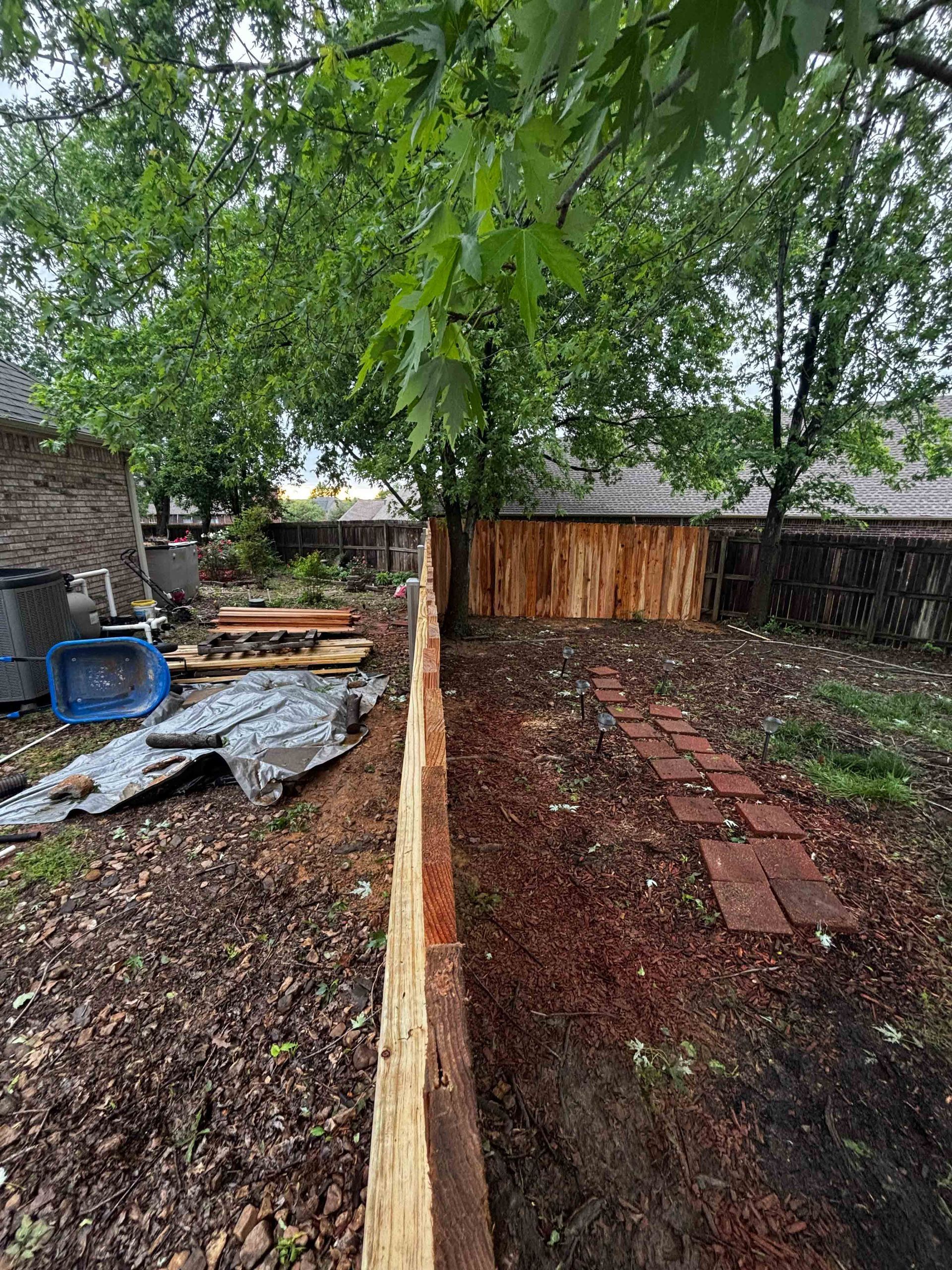 Backyard with a long wooden fence, wet ground, trees, and stacked materials after rain.