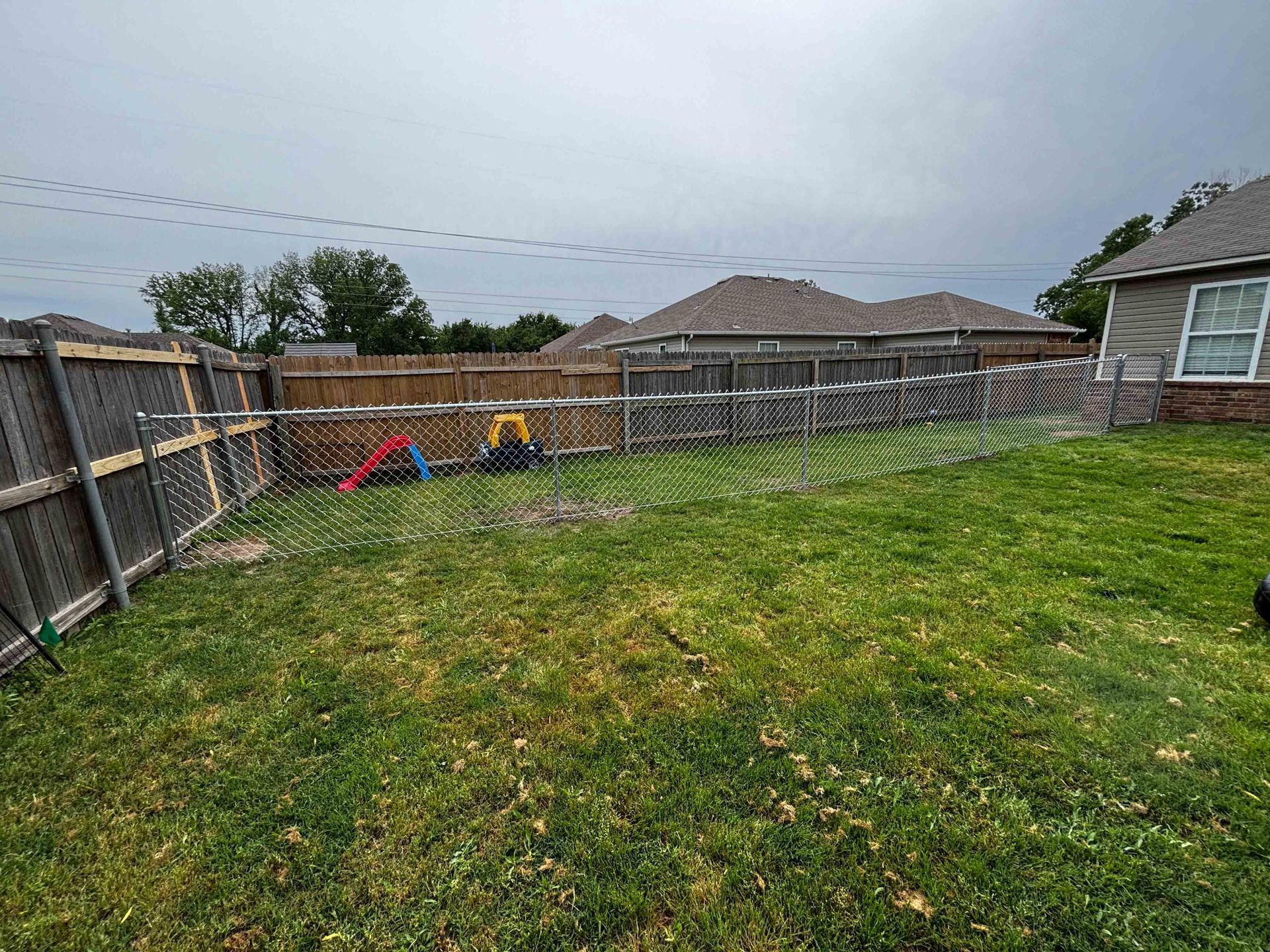 Backyard with green lawn, wooden fence, playset, and house under an overcast sky