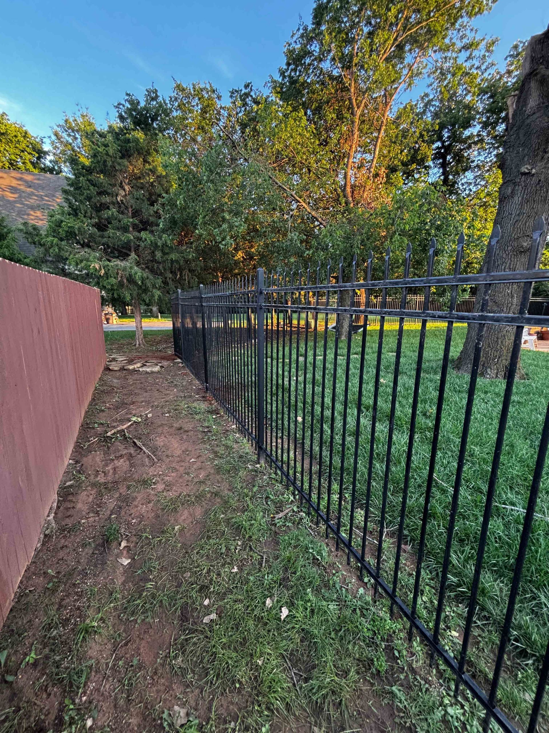 Narrow grassy path beside a black metal fence and red wall, with trees and a yard at sunset.