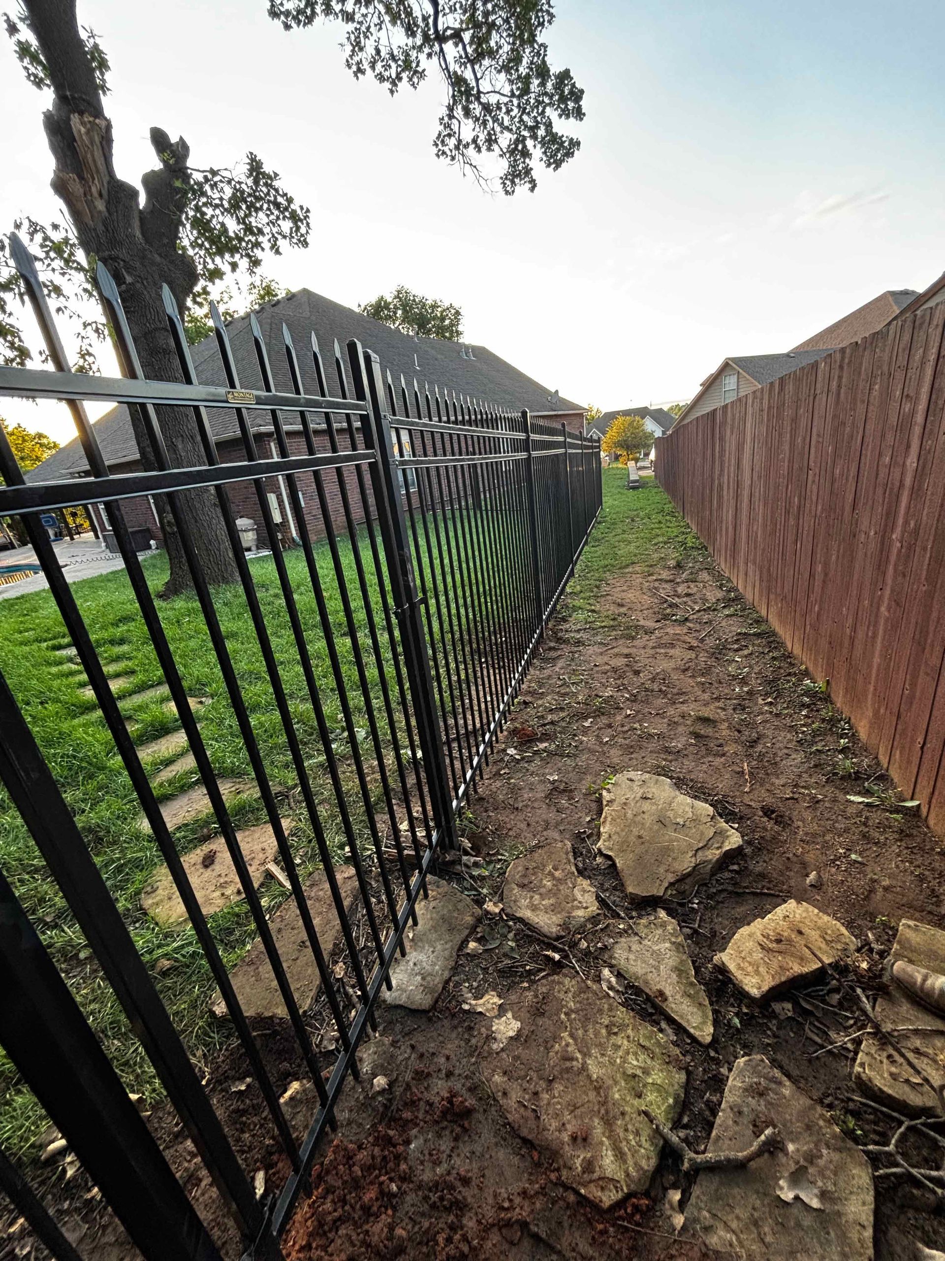 Narrow rocky path between a black metal fence and a brick wall, with grass and trees beside it.