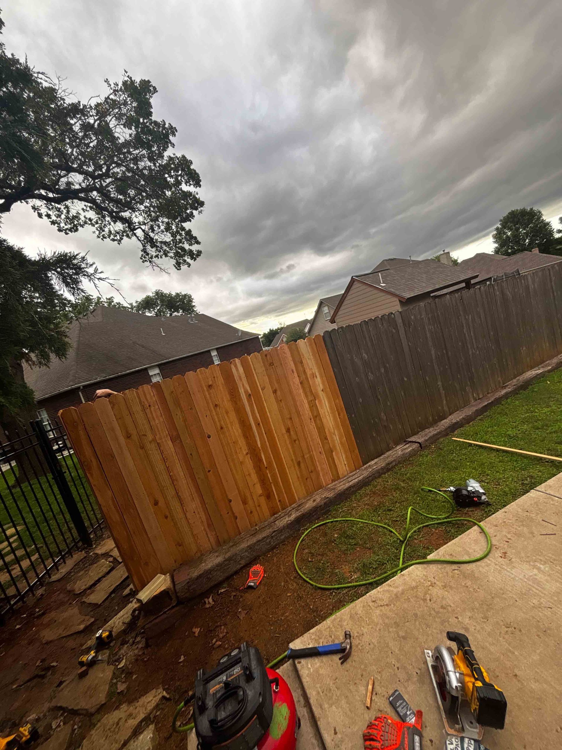 Backyard with a fallen wooden fence under stormy clouds, tools scattered on a wet concrete patio.