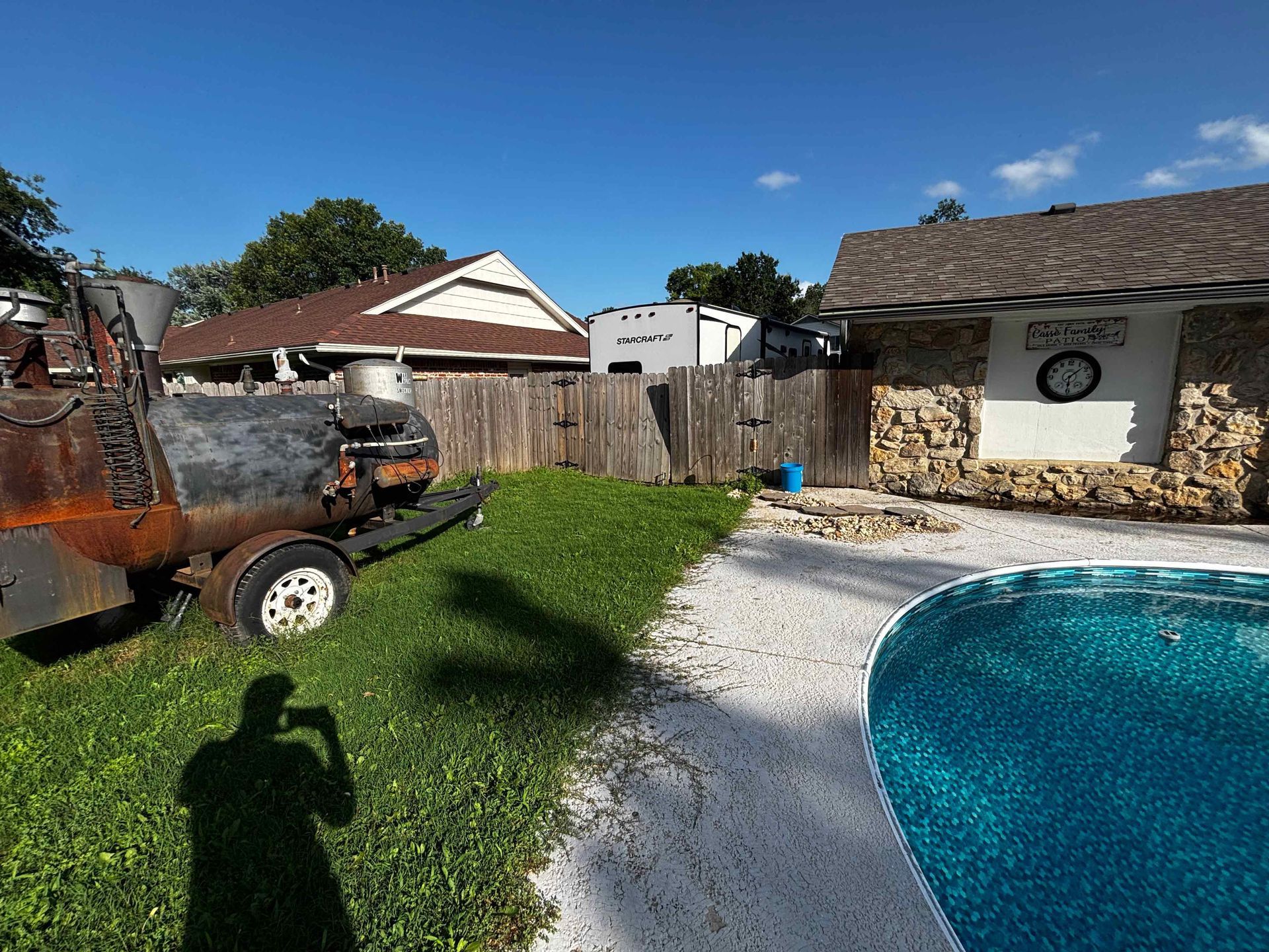 Backyard with swimming pool, lawn, brick grill area, and a large rusted trailer on the grass.