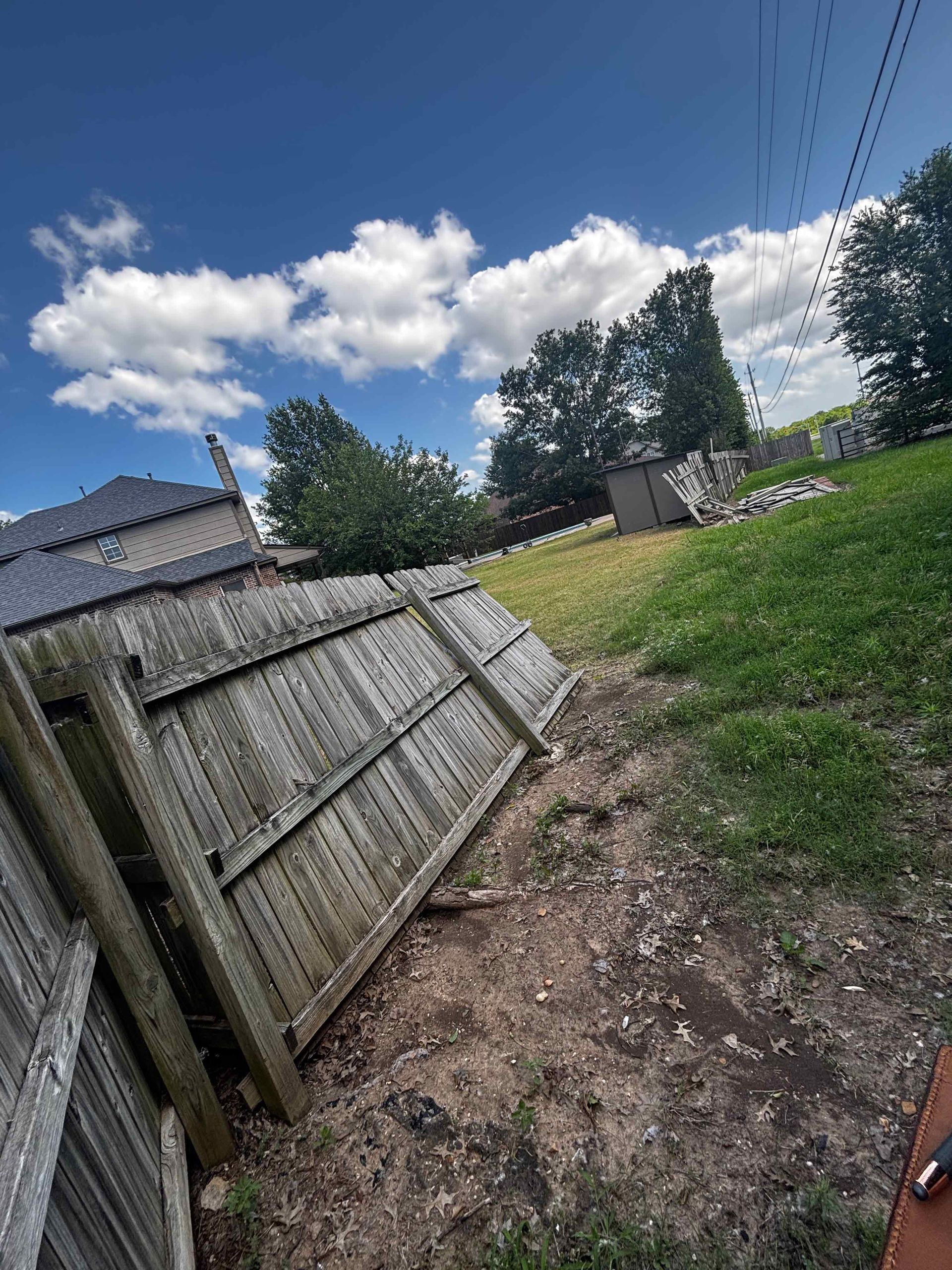 Tilted wooden fence beside a dirt path and grassy yard under a blue sky with clouds