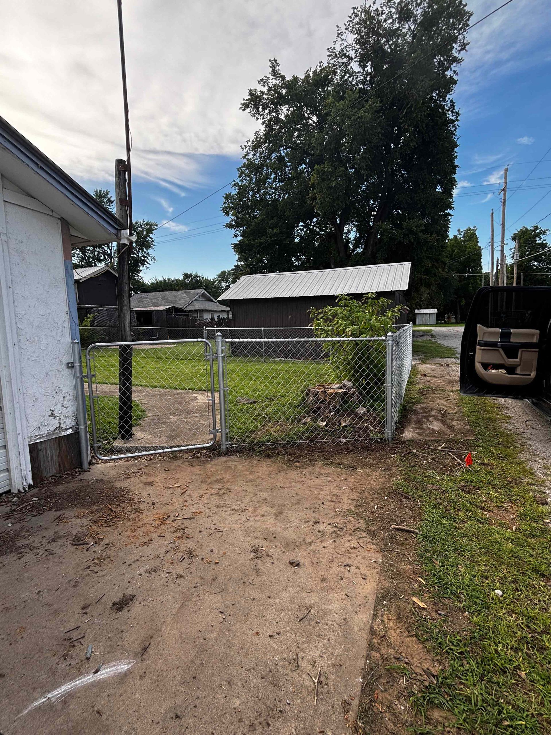 Open chain-link gate leading to a grassy yard with sheds and a large tree