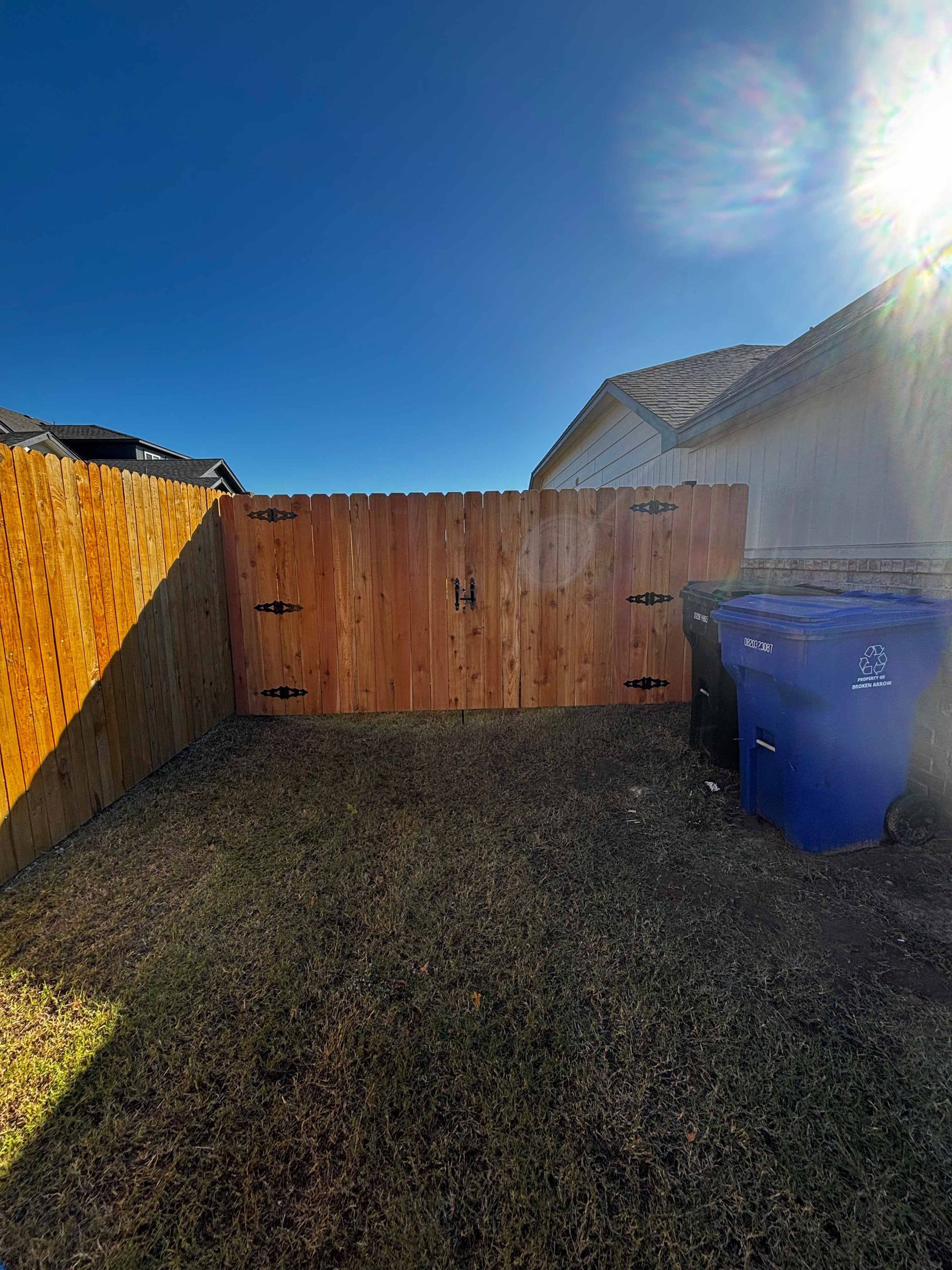 Sunlit backyard with wooden fence, gravel ground, and a blue trash bin beside a house.