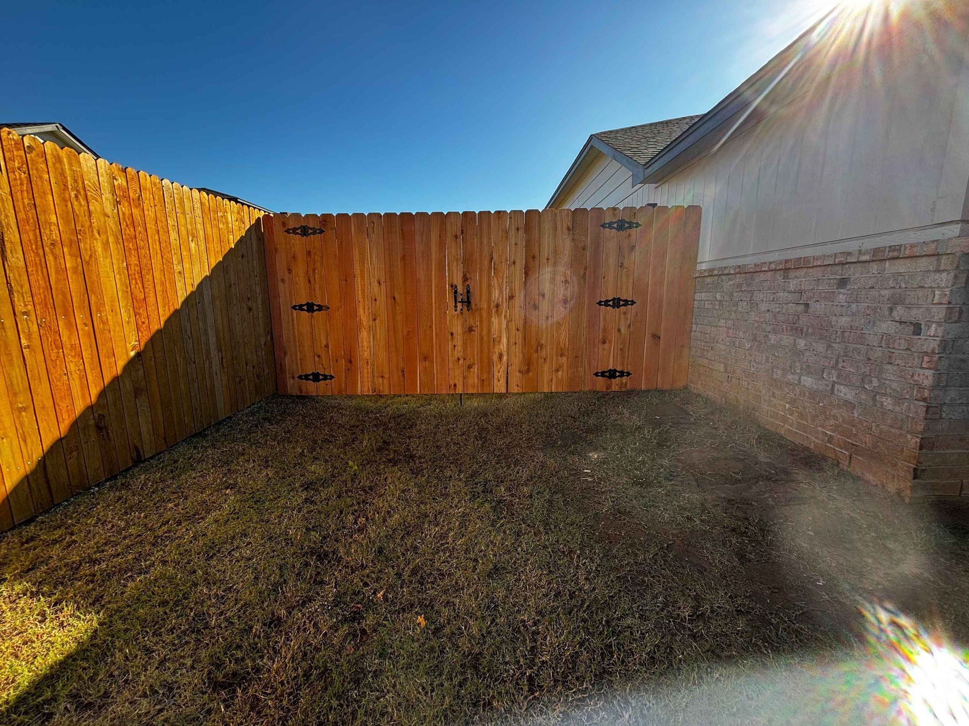 Sunlit backyard with a wooden fence and gate, patchy grass, and a house wall on the right.