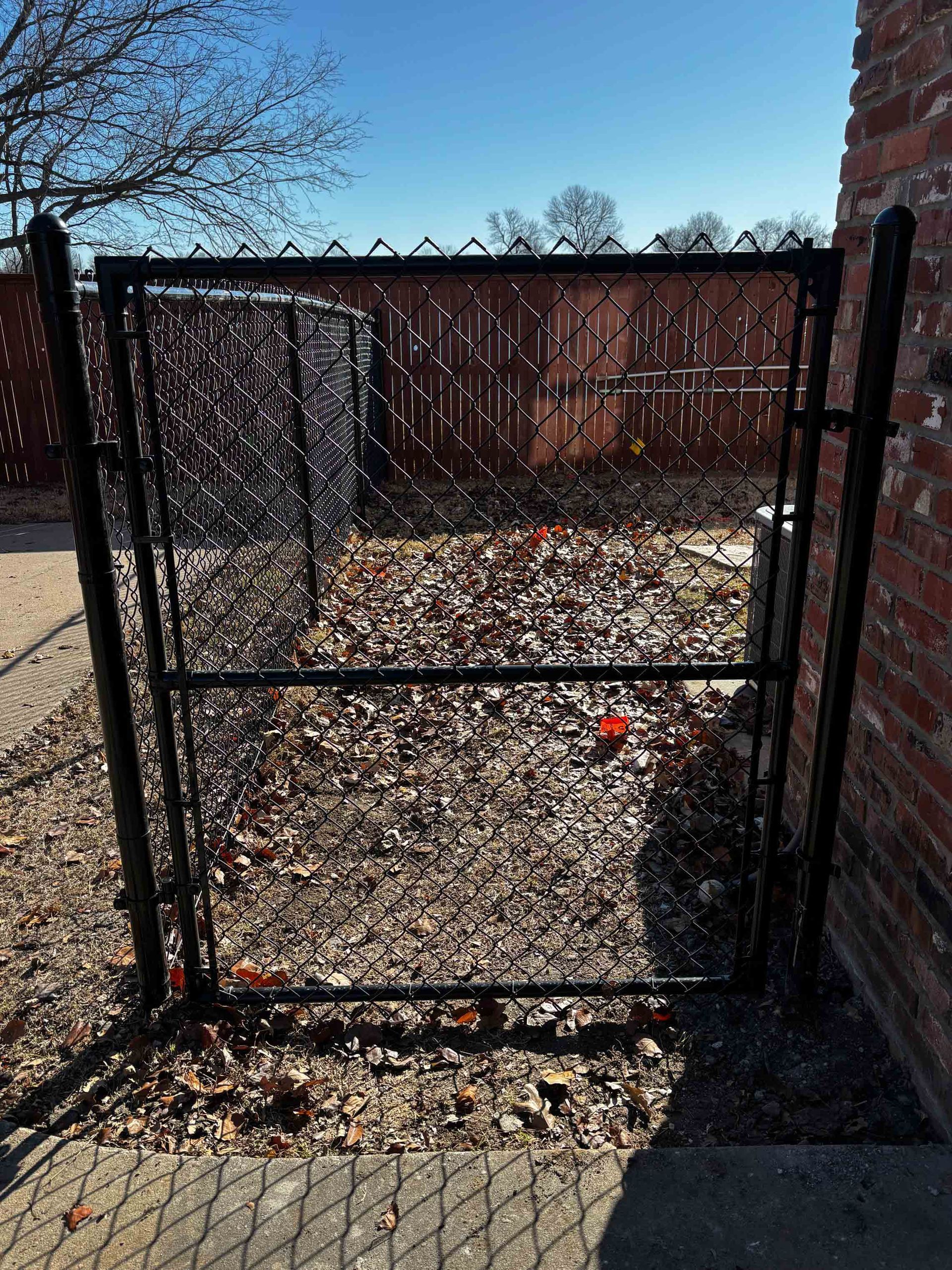 Black metal gate in a leaf-covered backyard beside a brick wall under a clear sky