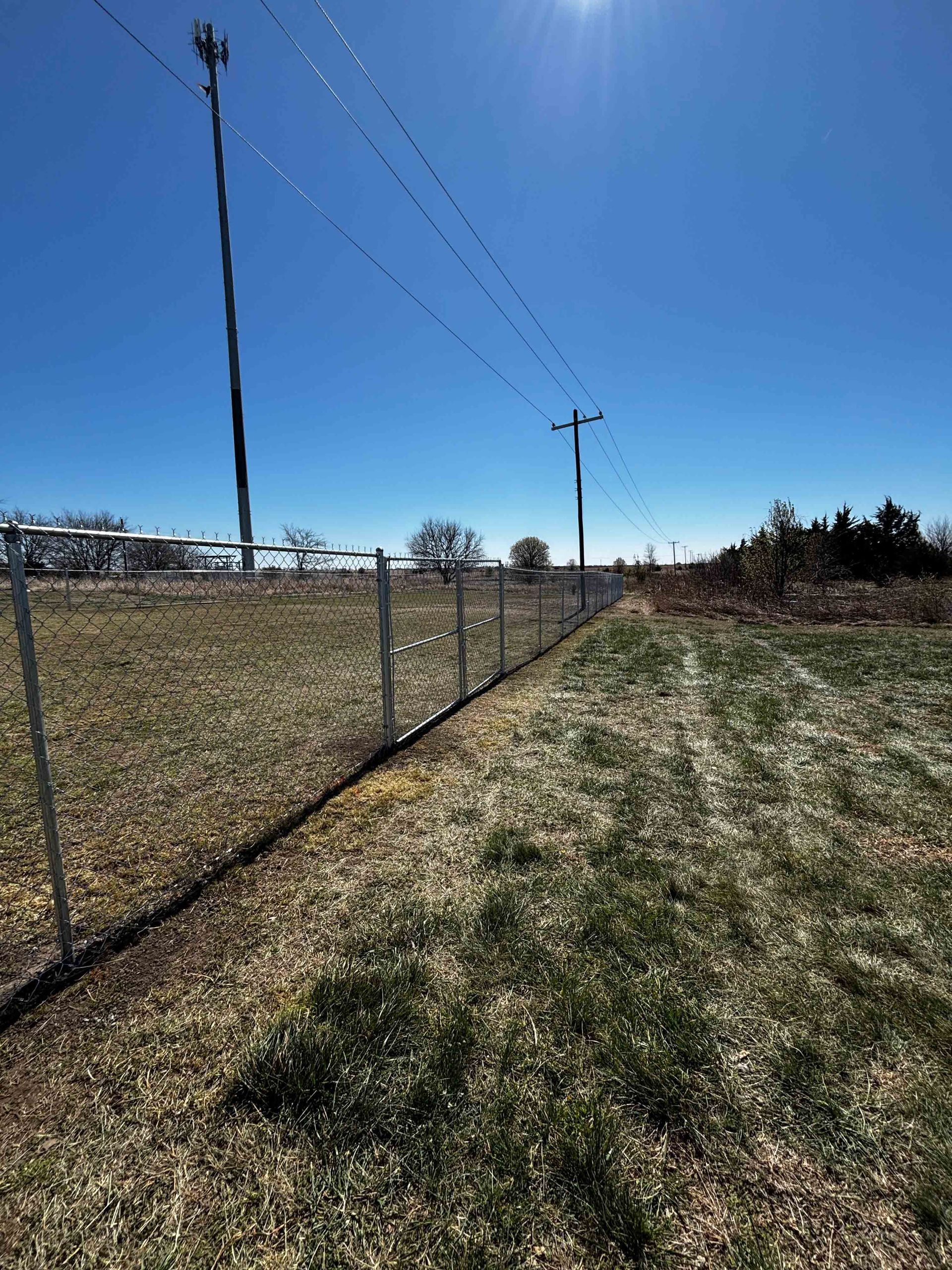 Chain-link fence beside grassy field under a clear blue sky with utility poles and power lines