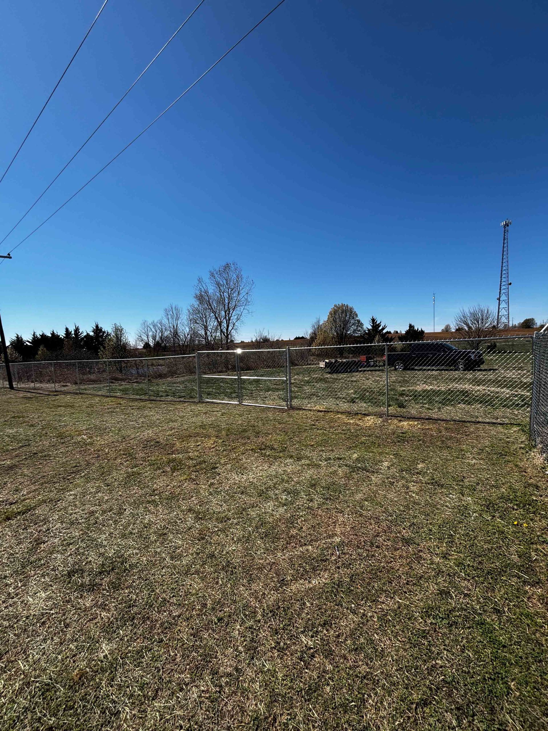 Open grassy field under a clear blue sky, with trees, power lines, and a distant tower.