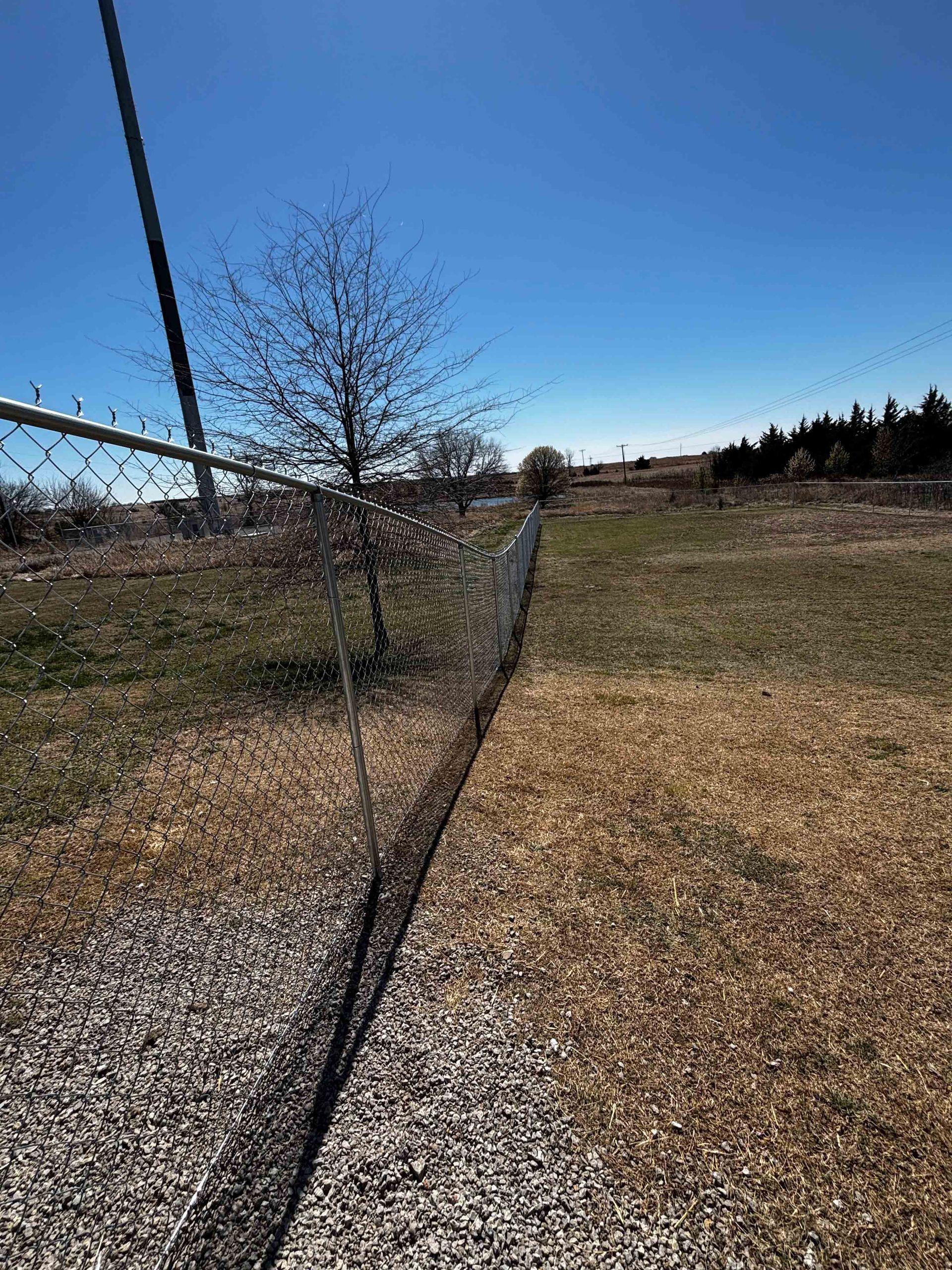Black netting fence runs along a gravel path beside a grassy field under a clear blue sky.