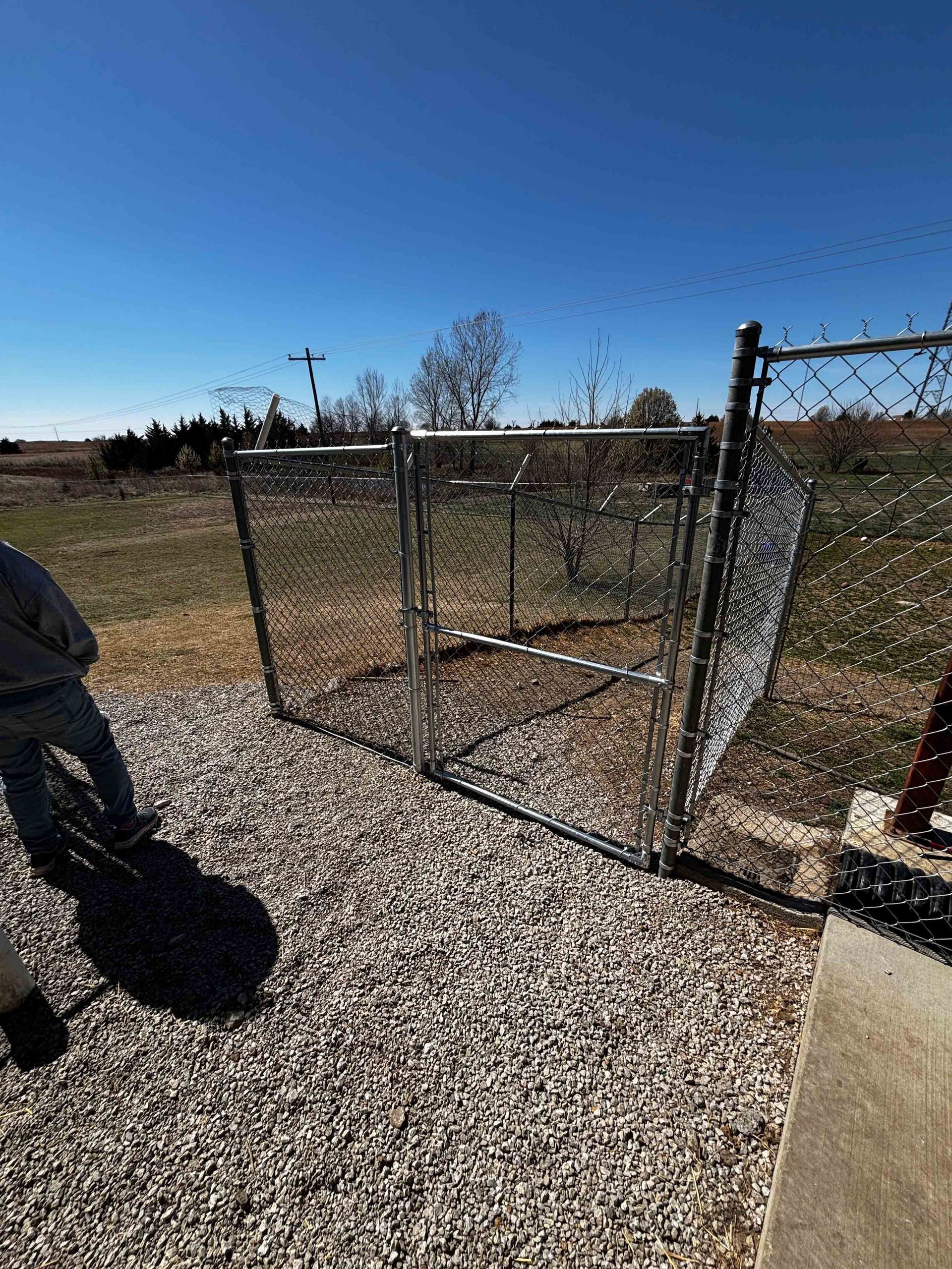 Chain-link kennel on gravel under a clear blue sky, with a person partially visible at left.